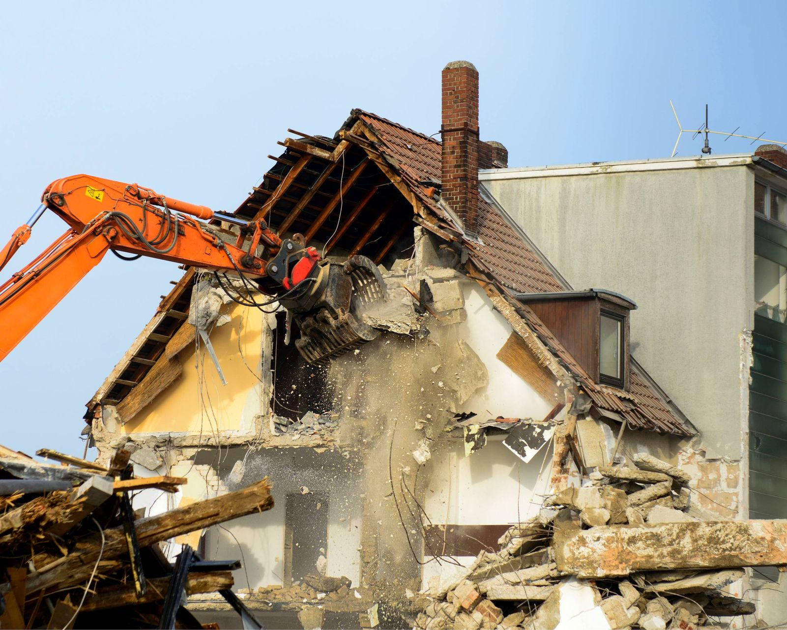 Orange excavator demolishing a house, with debris and dust flying against a blue sky.