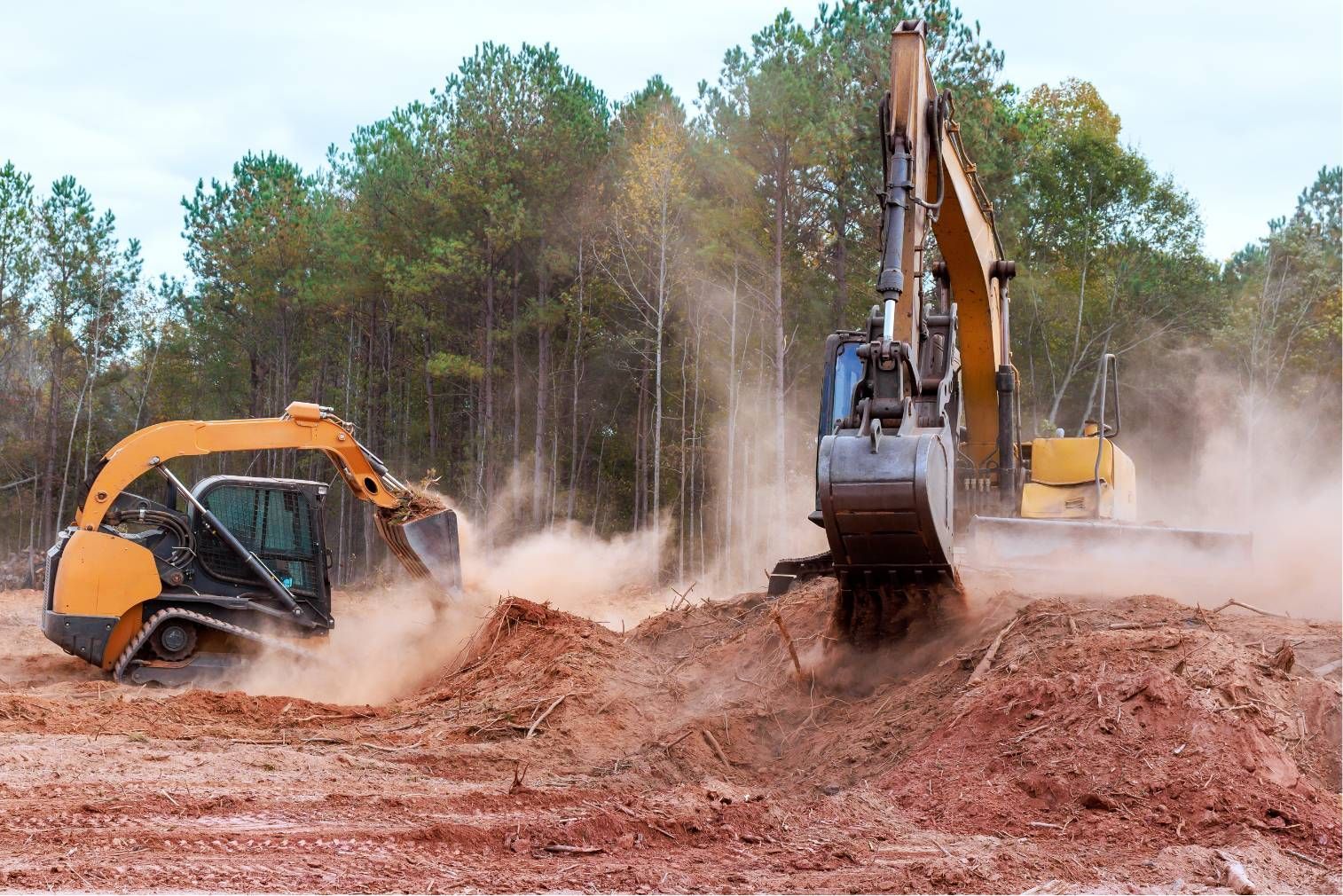 Excavator and orange dump truck kicking up dust at a muddy construction site with trees in the background