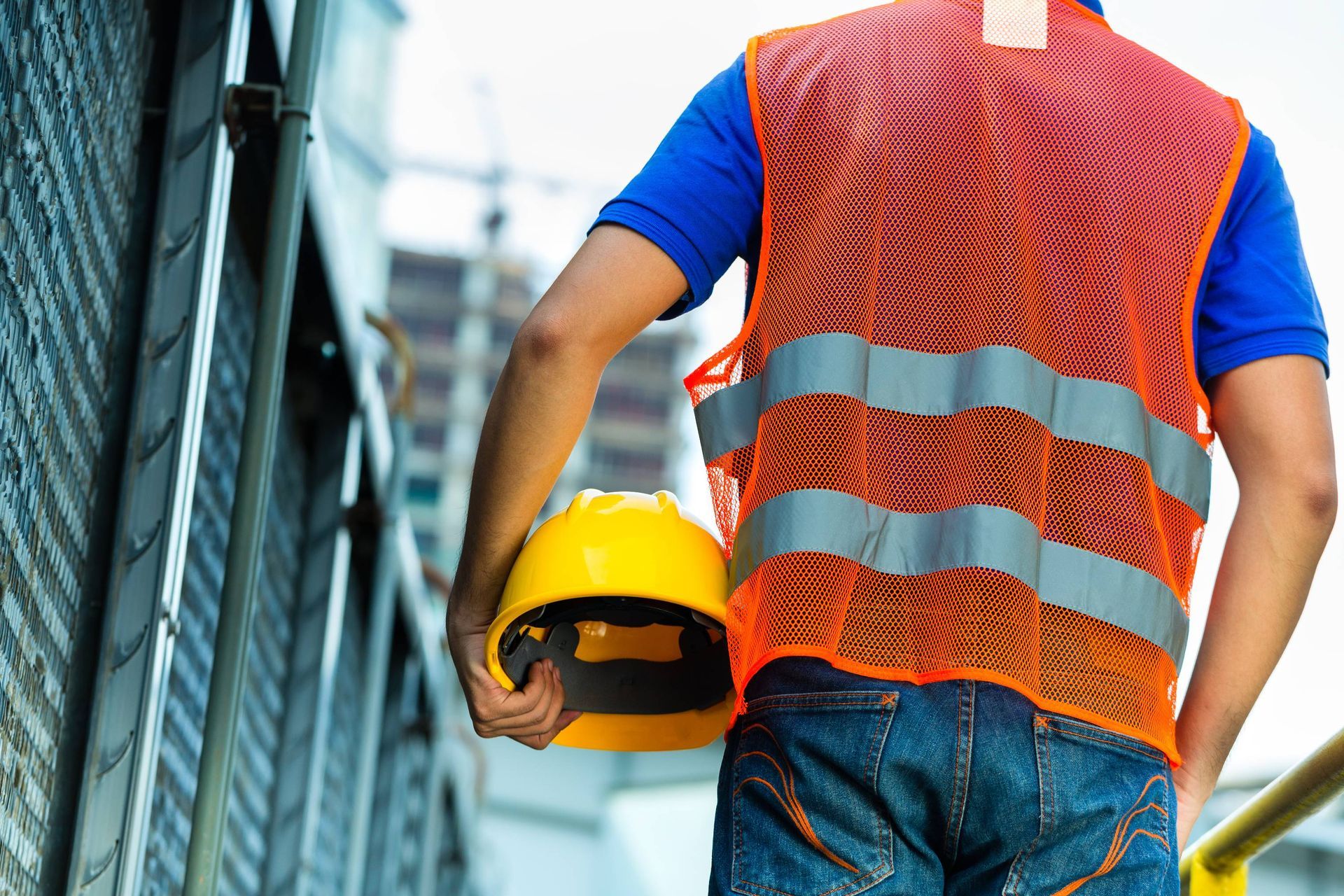 Construction worker in orange safety vest and yellow hard hat standing on a site, seen from behind.
