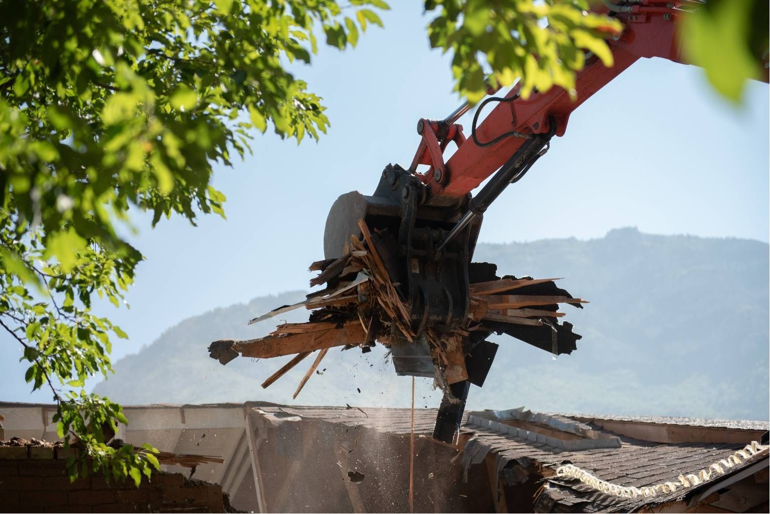 Excavator demolition claw tearing down a roof with mountains in the background