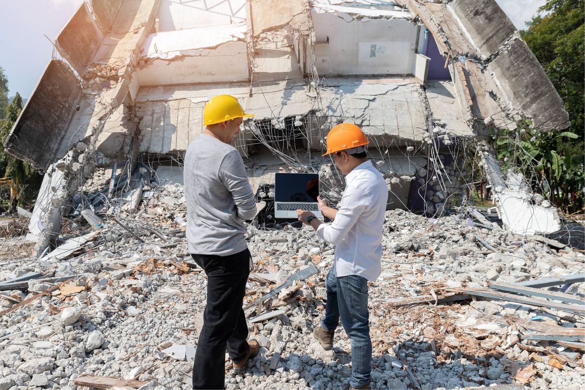 Two workers in hard hats inspect earthquake rubble before a collapsed building