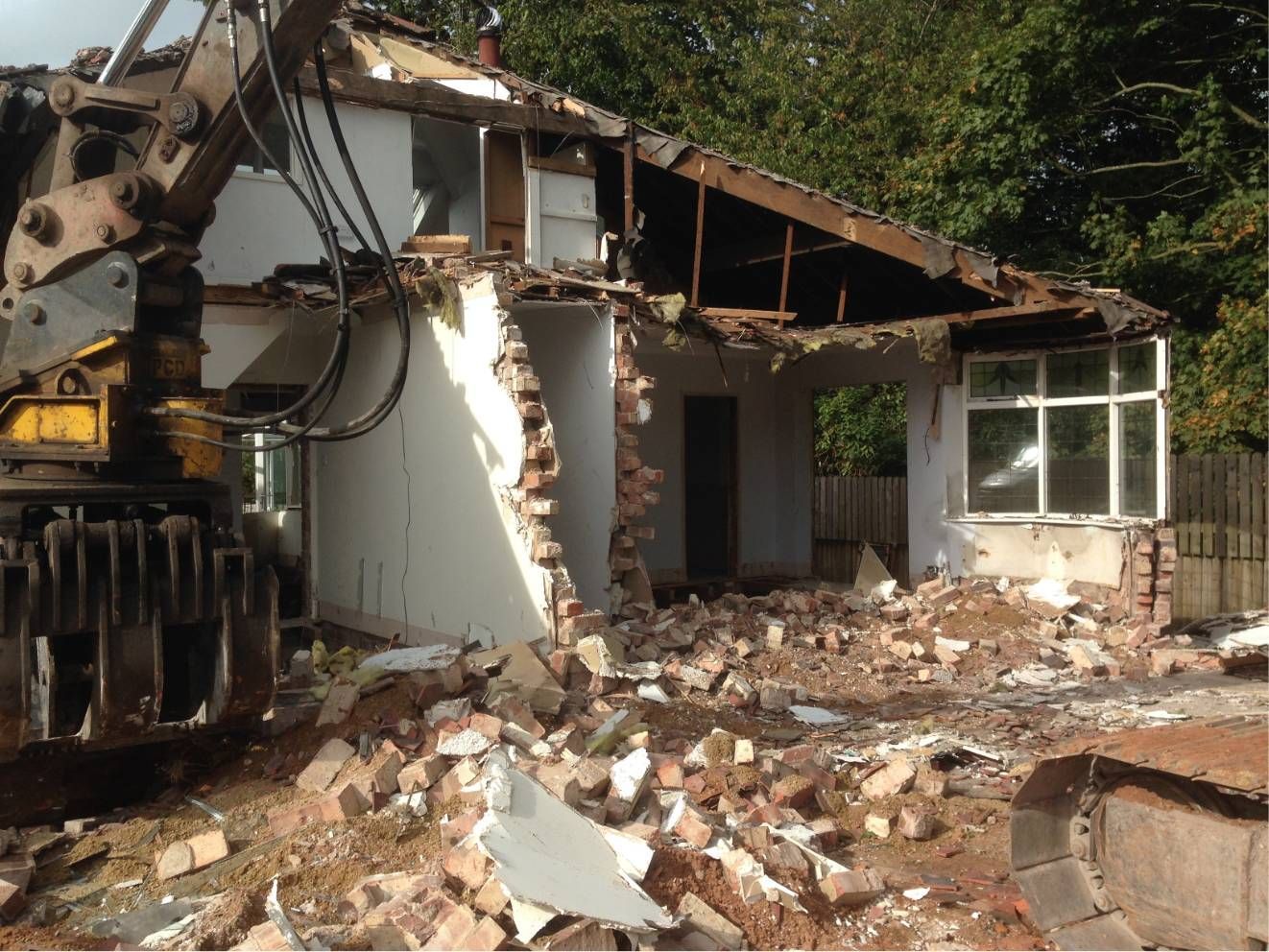 Excavator demolishing a damaged brick house, with rubble and debris in the foreground.
