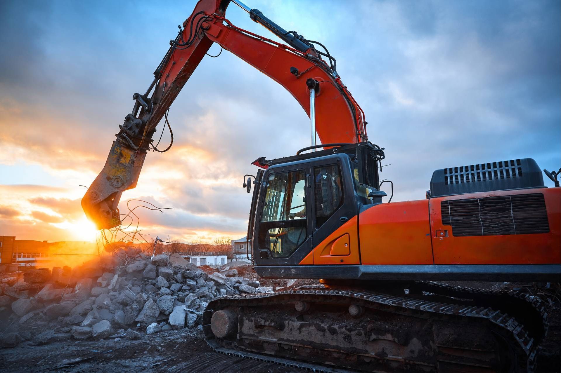 Orange excavator on a rocky construction site at sunset, digging rubble under a dramatic sky