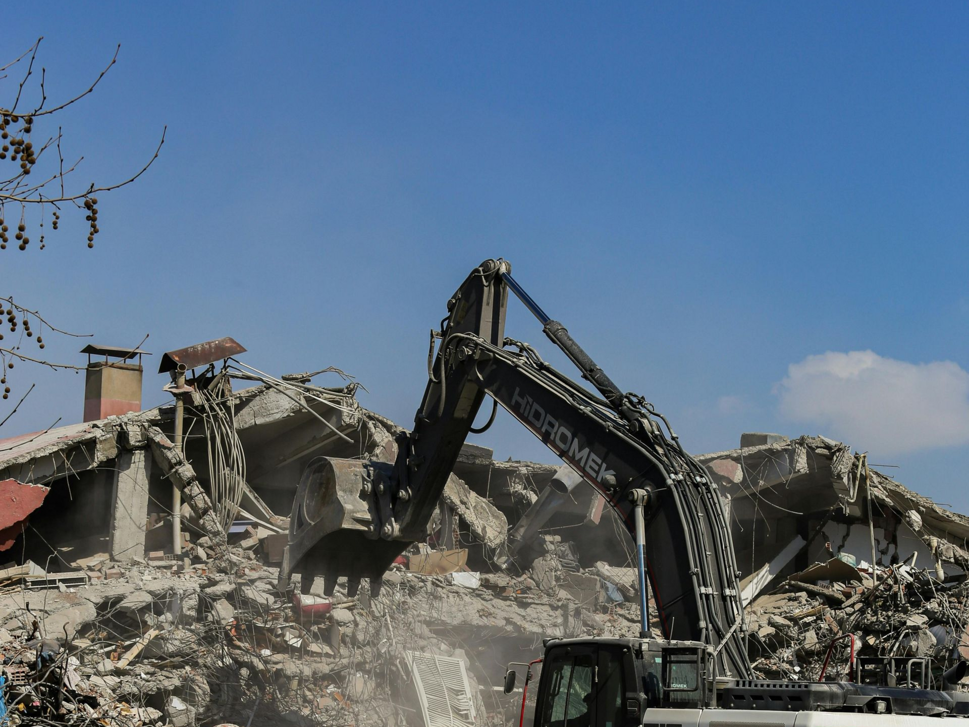 Excavator demolishing a collapsed building amid rubble under a clear blue sky