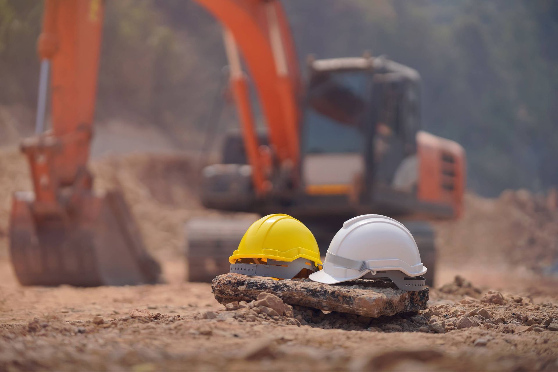 Two hard hats on dirt at a construction site with an excavator in the background