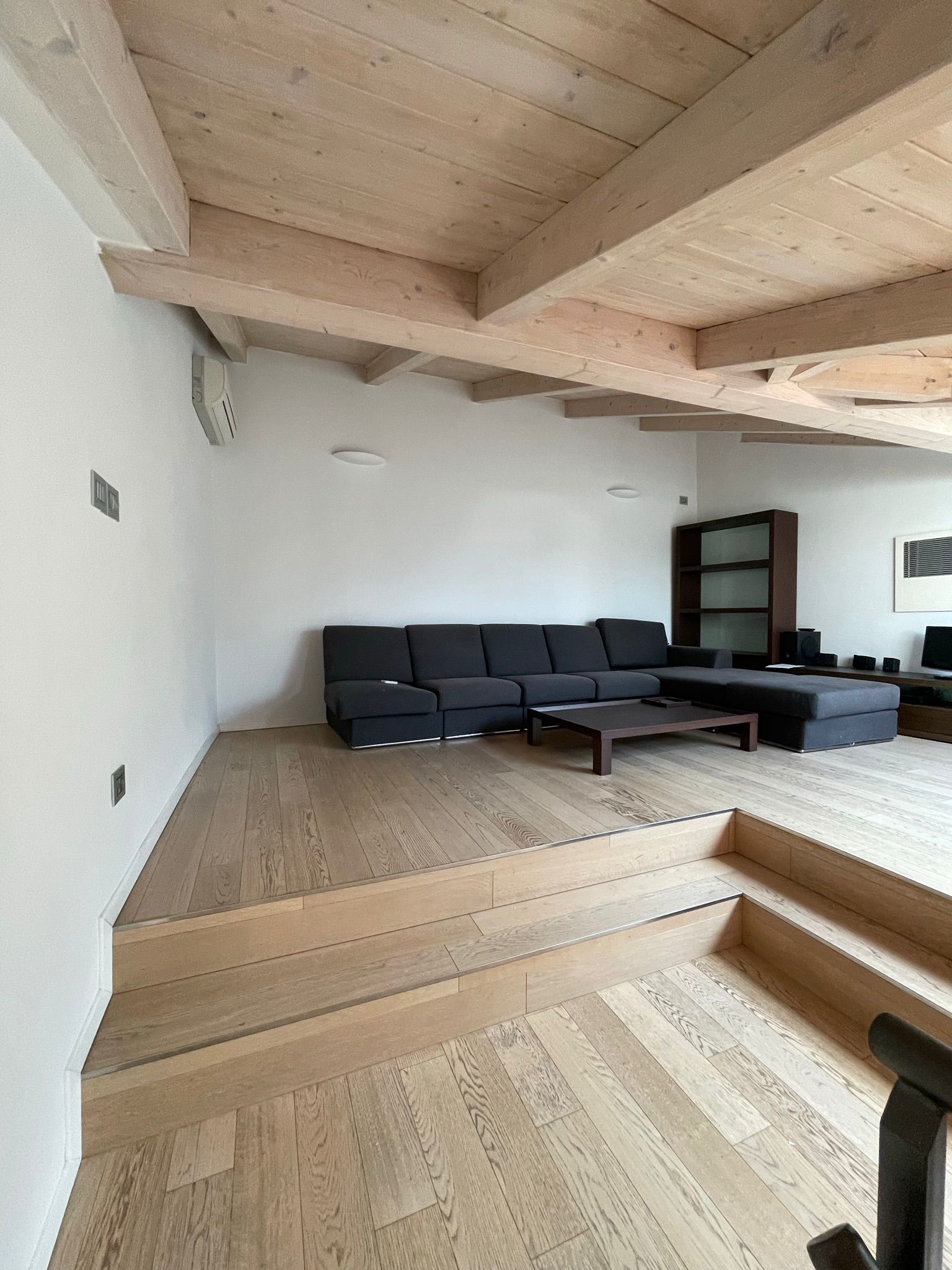 Una zona giorno con un divano scuro su una piattaforma di legno rialzata, caratterizzata da travi in legno chiaro e una scala a più livelli.