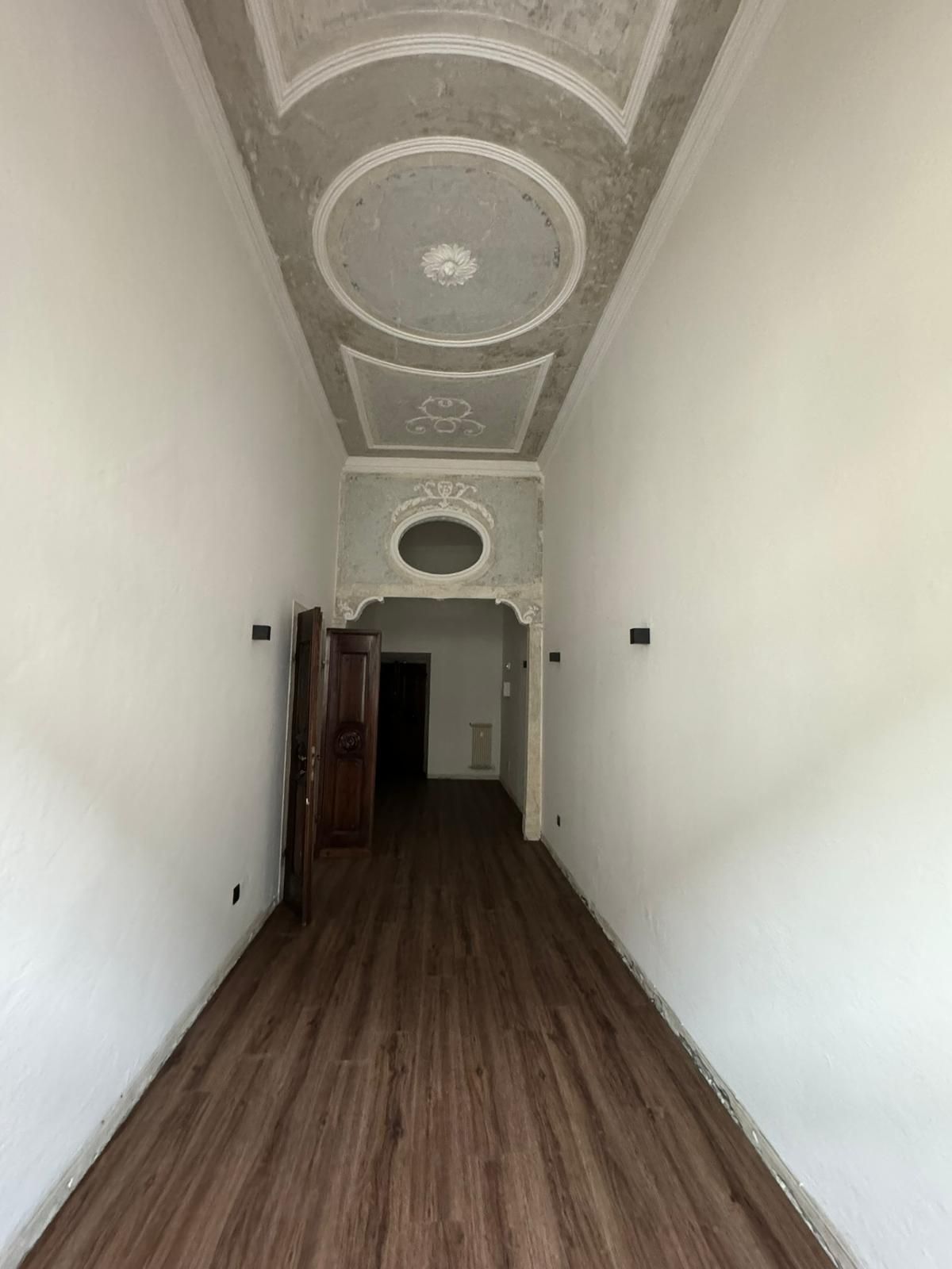 Un lungo corridoio con pareti bianche, pavimento effetto legno e un soffitto decorato e ornato con modanature circolari.