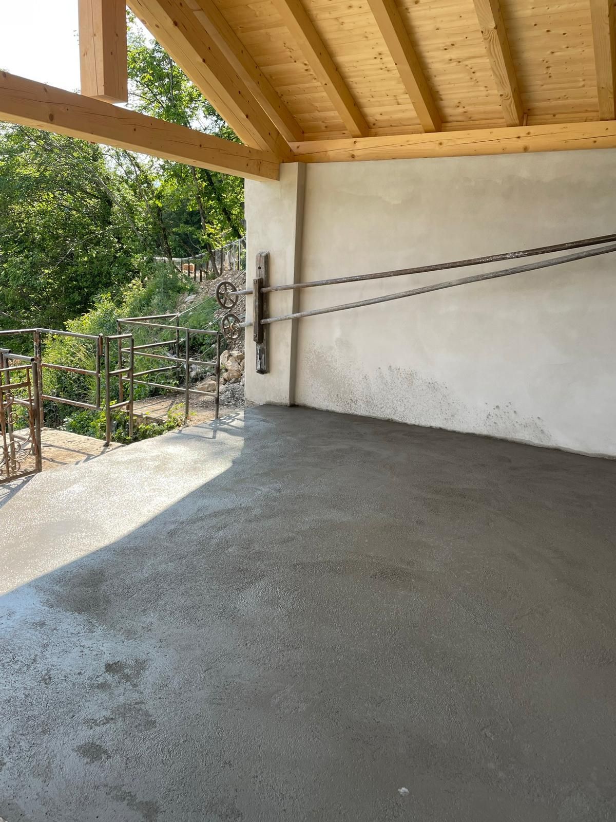 Un patio esterno coperto con pavimento in cemento grigio appena gettato, tetto con travi in legno e parete beige con cavi d'acciaio.