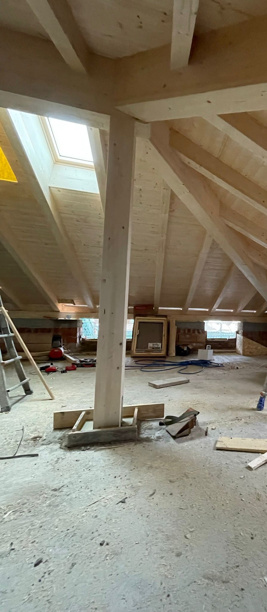 Una mansarda in costruzione con un pilastro centrale di sostegno in legno, soffitto con travi a vista e lucernari.