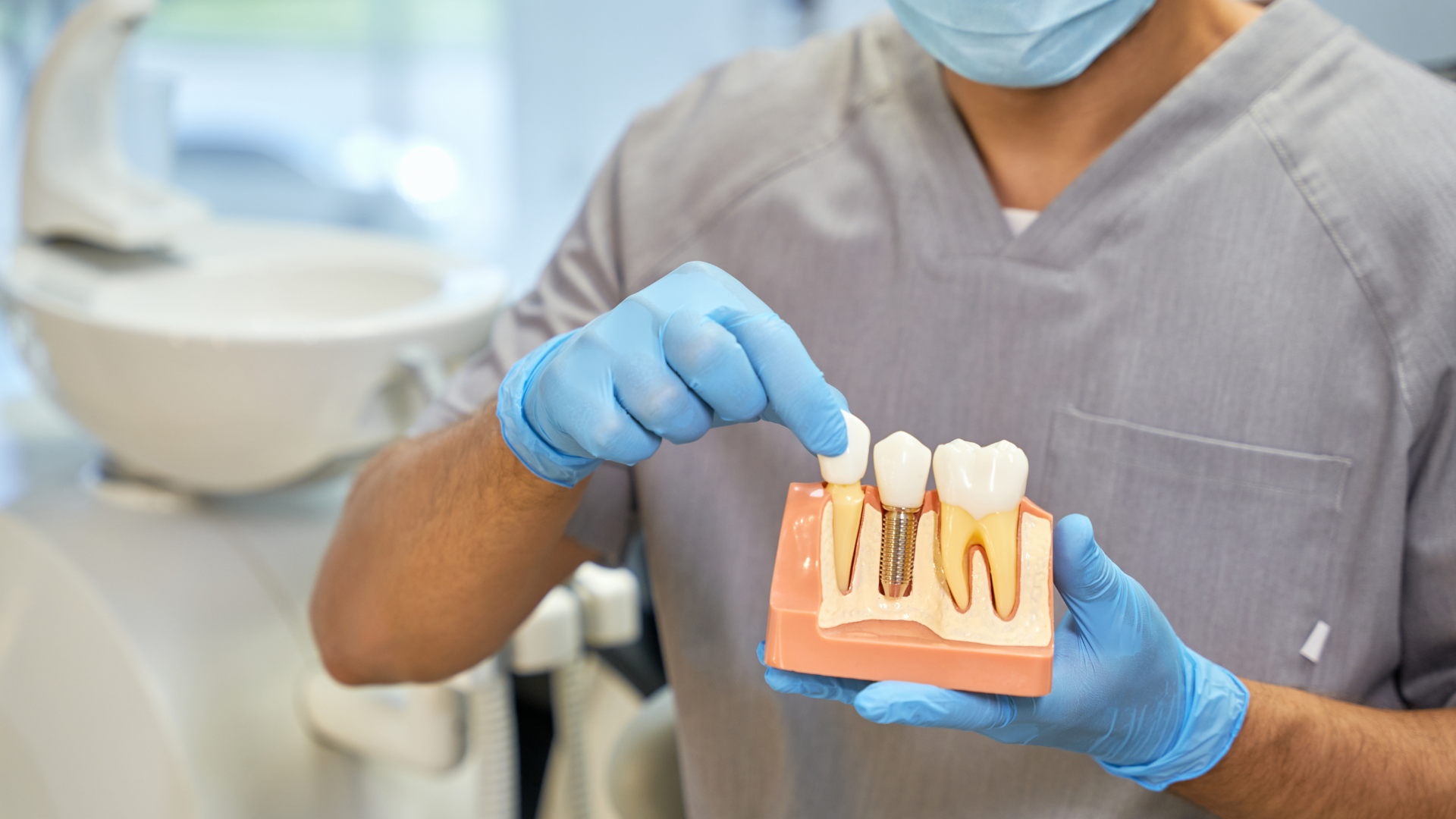Dentist in scrubs, mask, and gloves points at tooth model showing implant. Clinic setting.