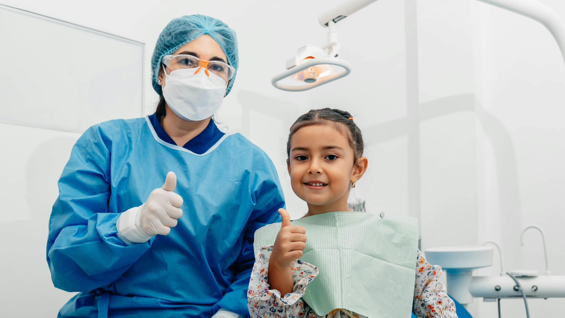 Dentist and child in a bright clinic, both giving thumbs up while the child wears a bib.