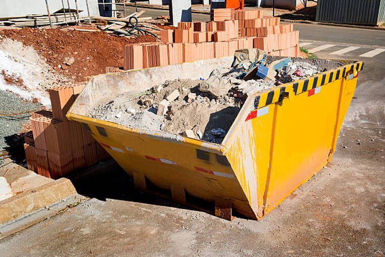 Yellow construction dumpster filled with debris; bricks in background.