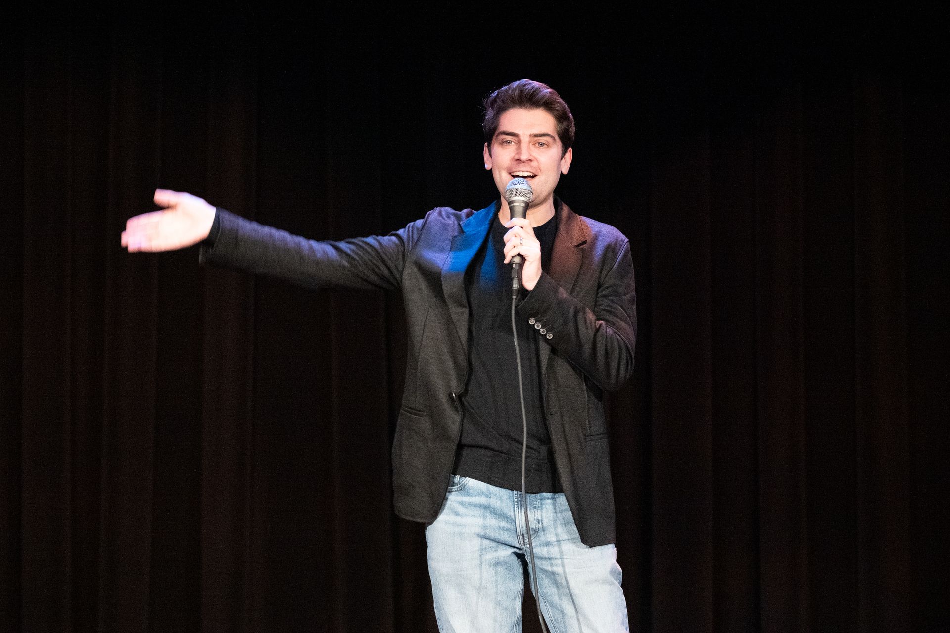 Man on stage holding a microphone, gesturing with his arm. Dark jacket, blue jeans. Black background.