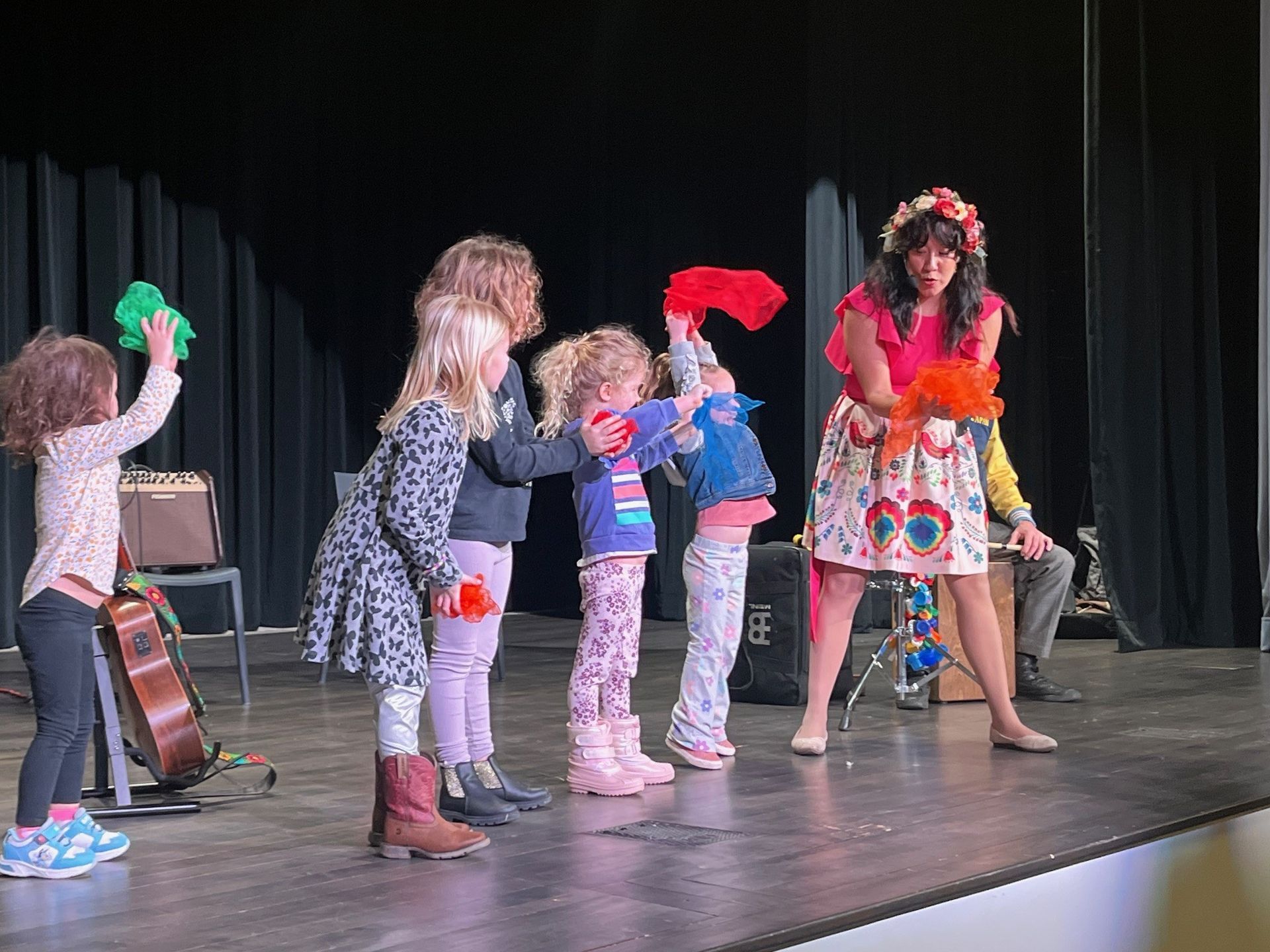 Children and an adult on a stage, holding up colorful fabrics. There is a guitar and amp on the left.