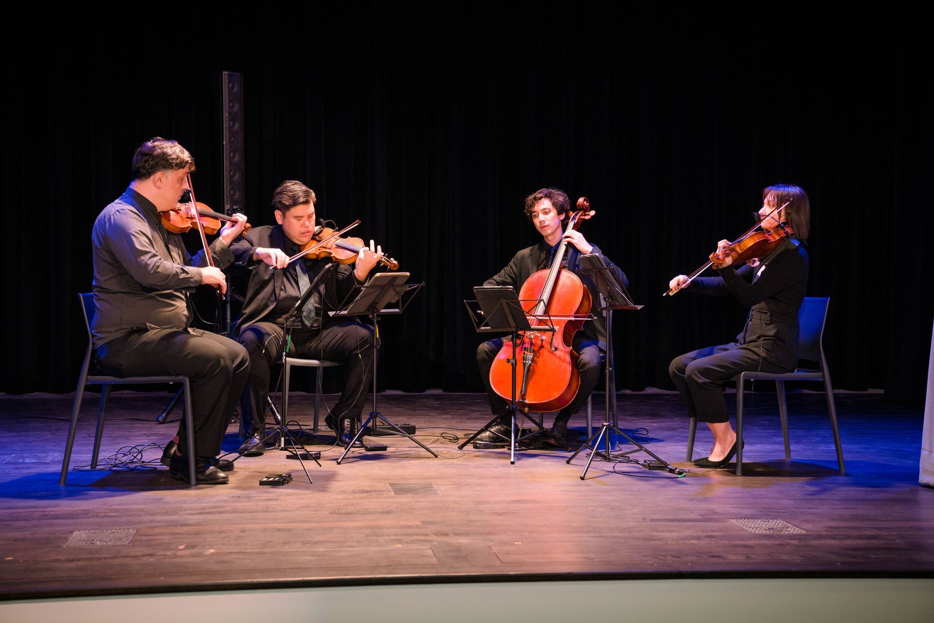 String quartet performing on stage, dark setting. Musicians playing instruments, music stands present.