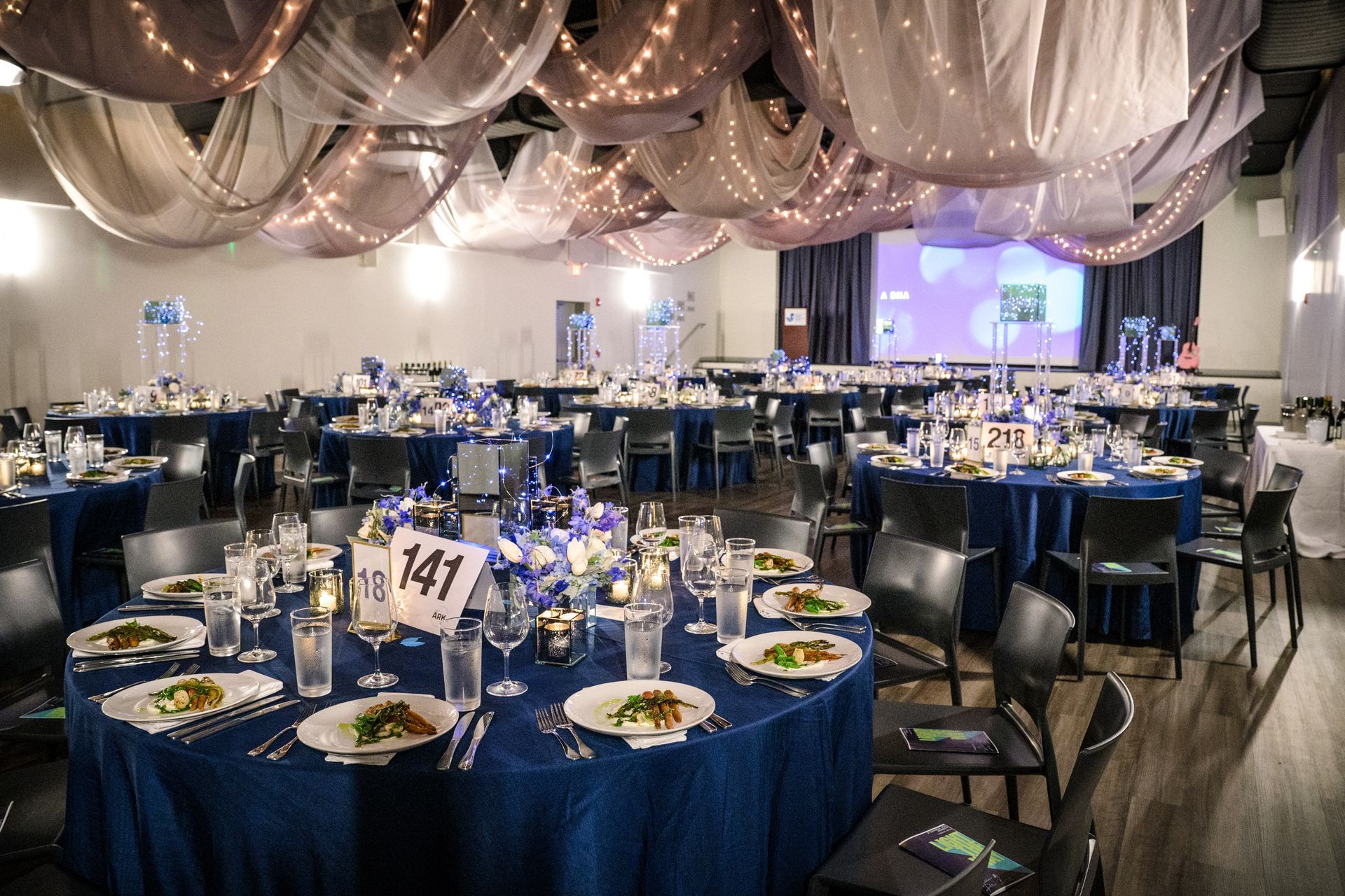 Elegant ballroom set for a dinner with blue tablecloths, centerpieces, and draping, with a stage in the background.
