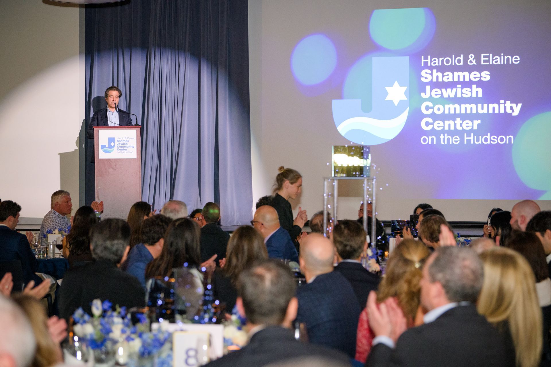 Man speaking at a podium in a Harold & Elaine Shames Jewish Community Center. Audience applauds in a banquet hall.