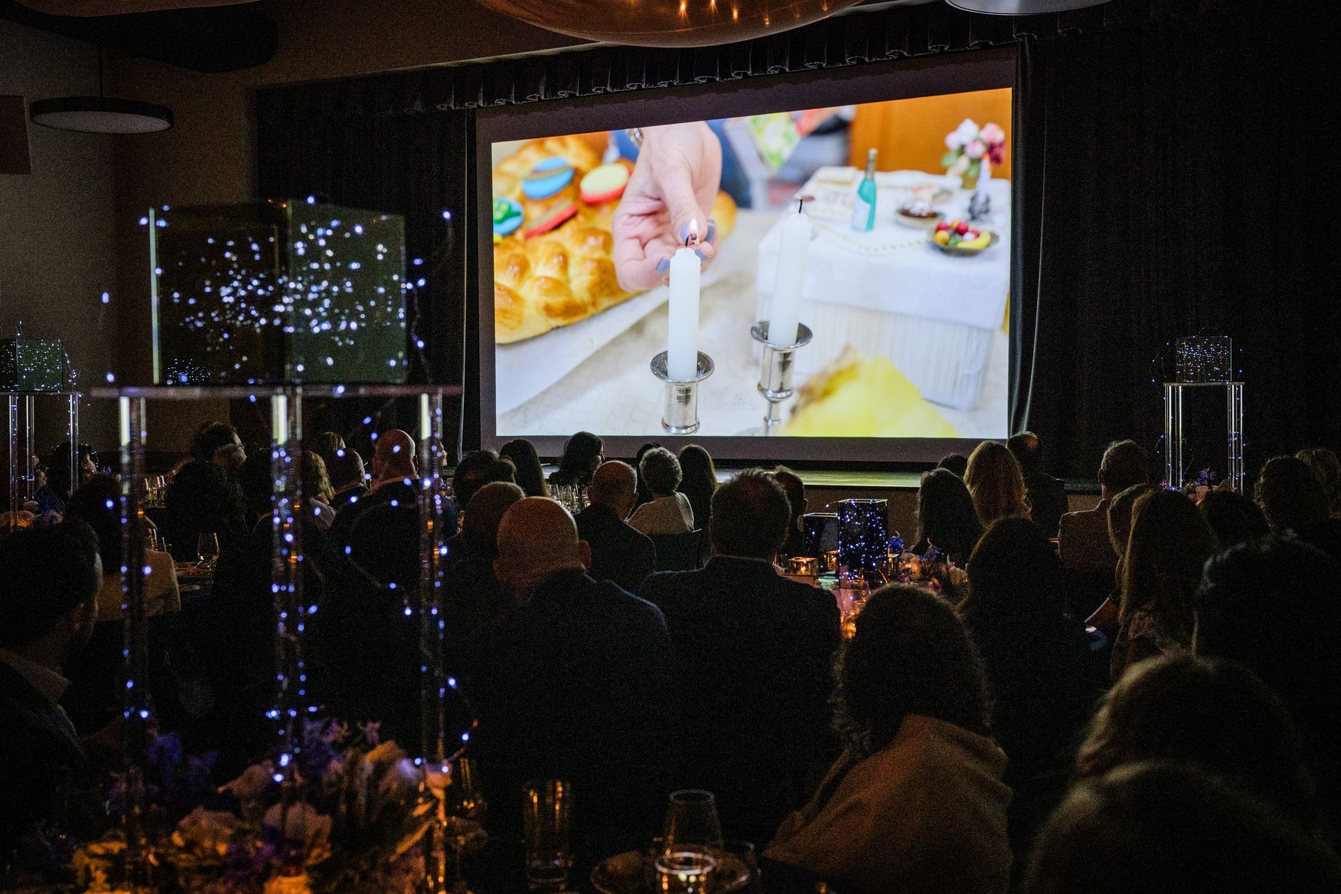 Audience watches screen at event, showing a close-up of a person lighting candles on a table.