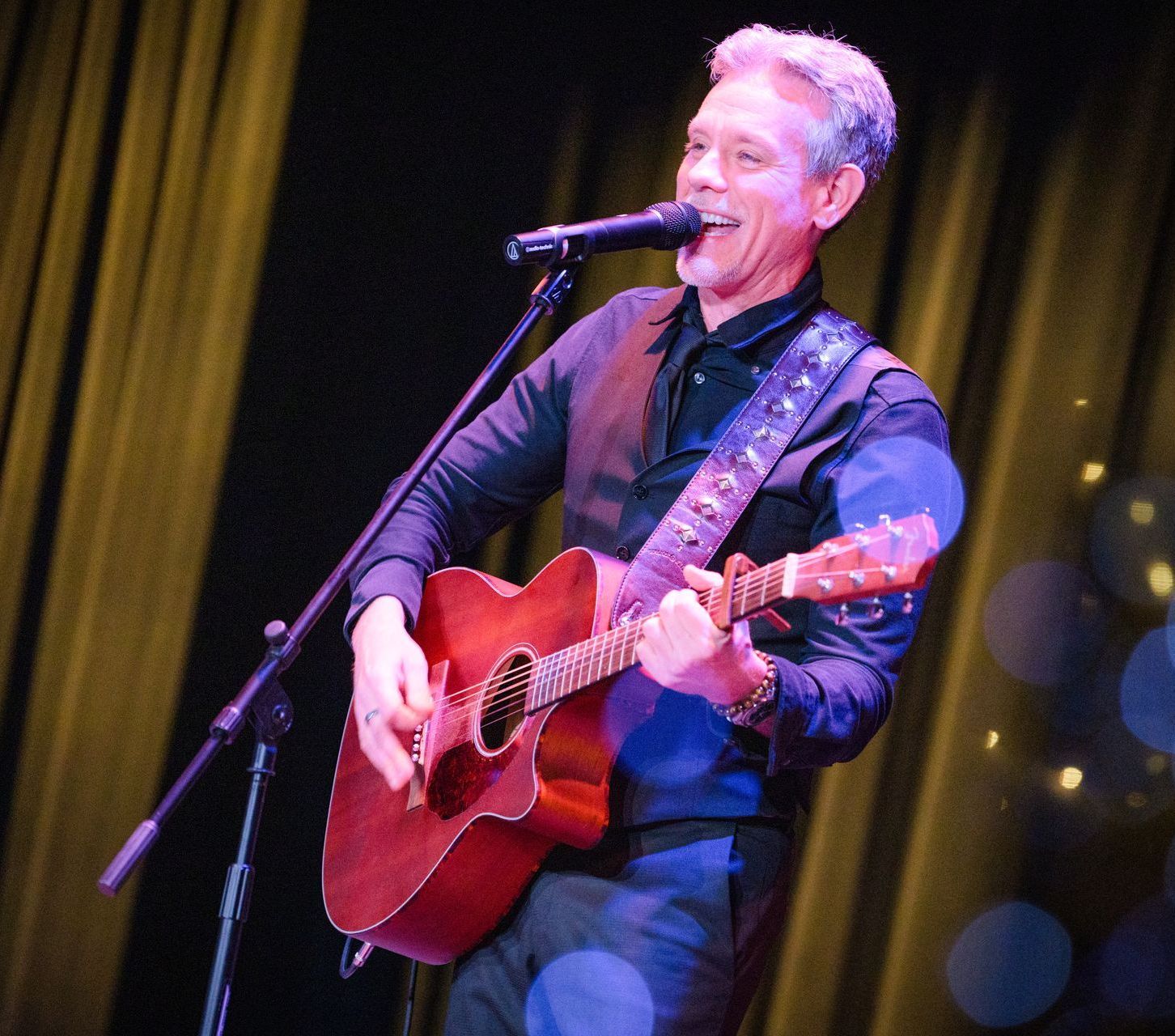 Man playing acoustic guitar on stage, singing into a microphone. Dark clothing, stage lights.