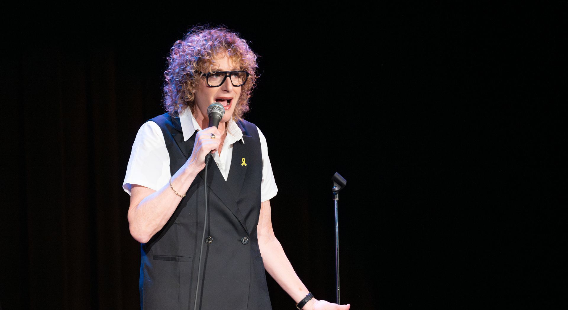 Comedian with curly hair wearing glasses, speaking into a microphone on stage, black backdrop.