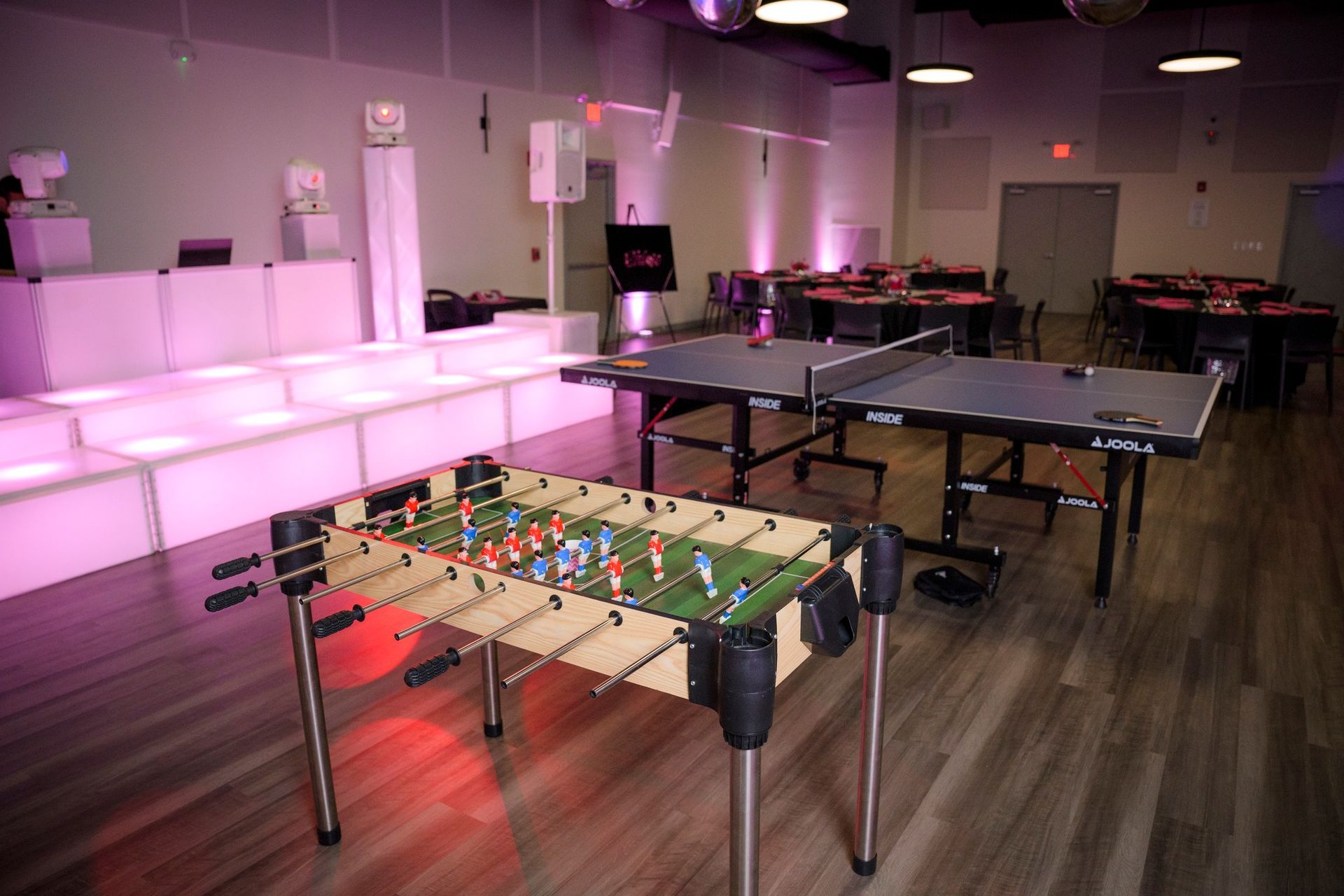 Foosball and ping pong tables in a lit party room with tables and a DJ setup.