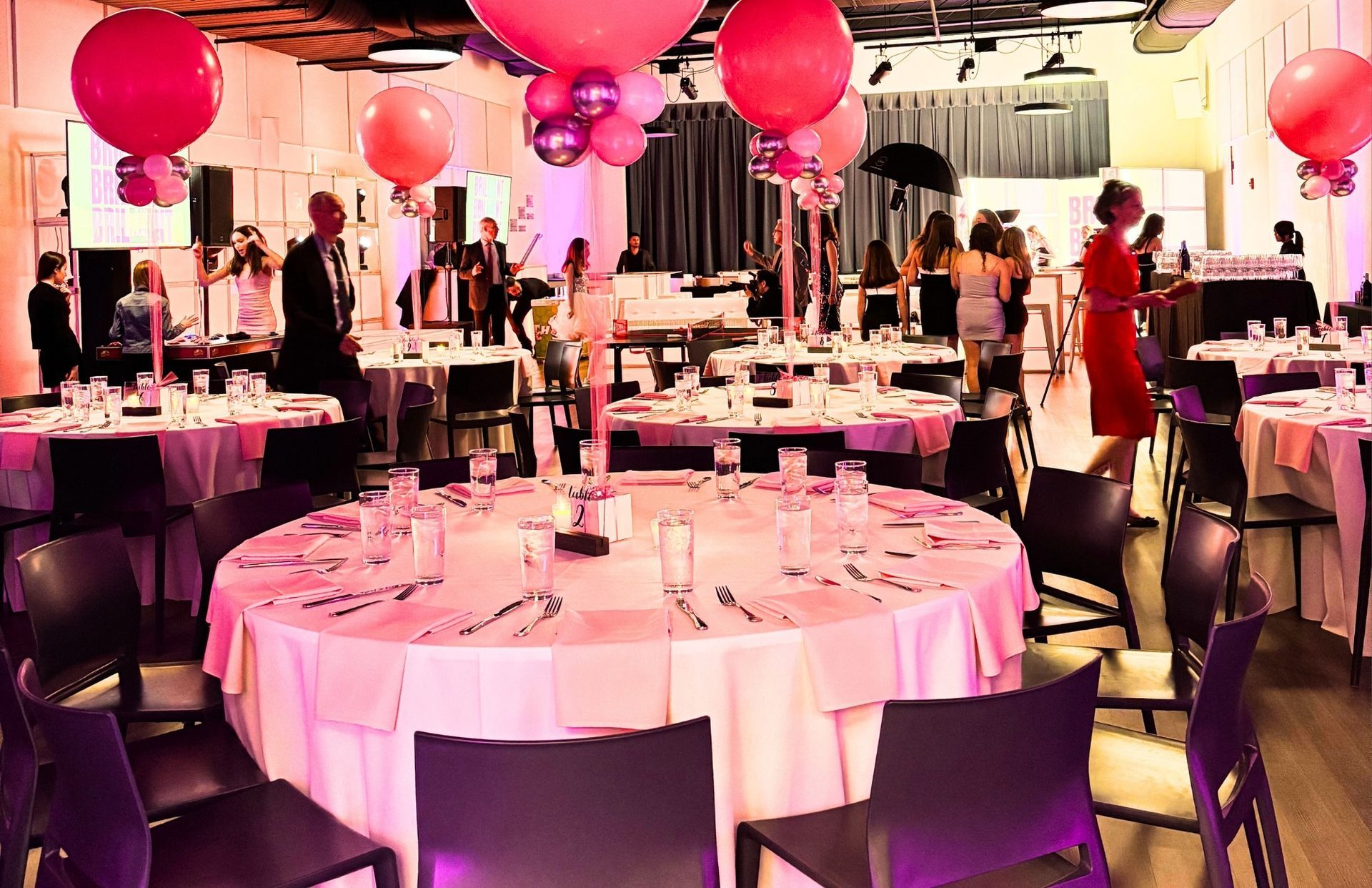 Pink-themed event with round tables set for dining. Large pink balloons and people mingling.
