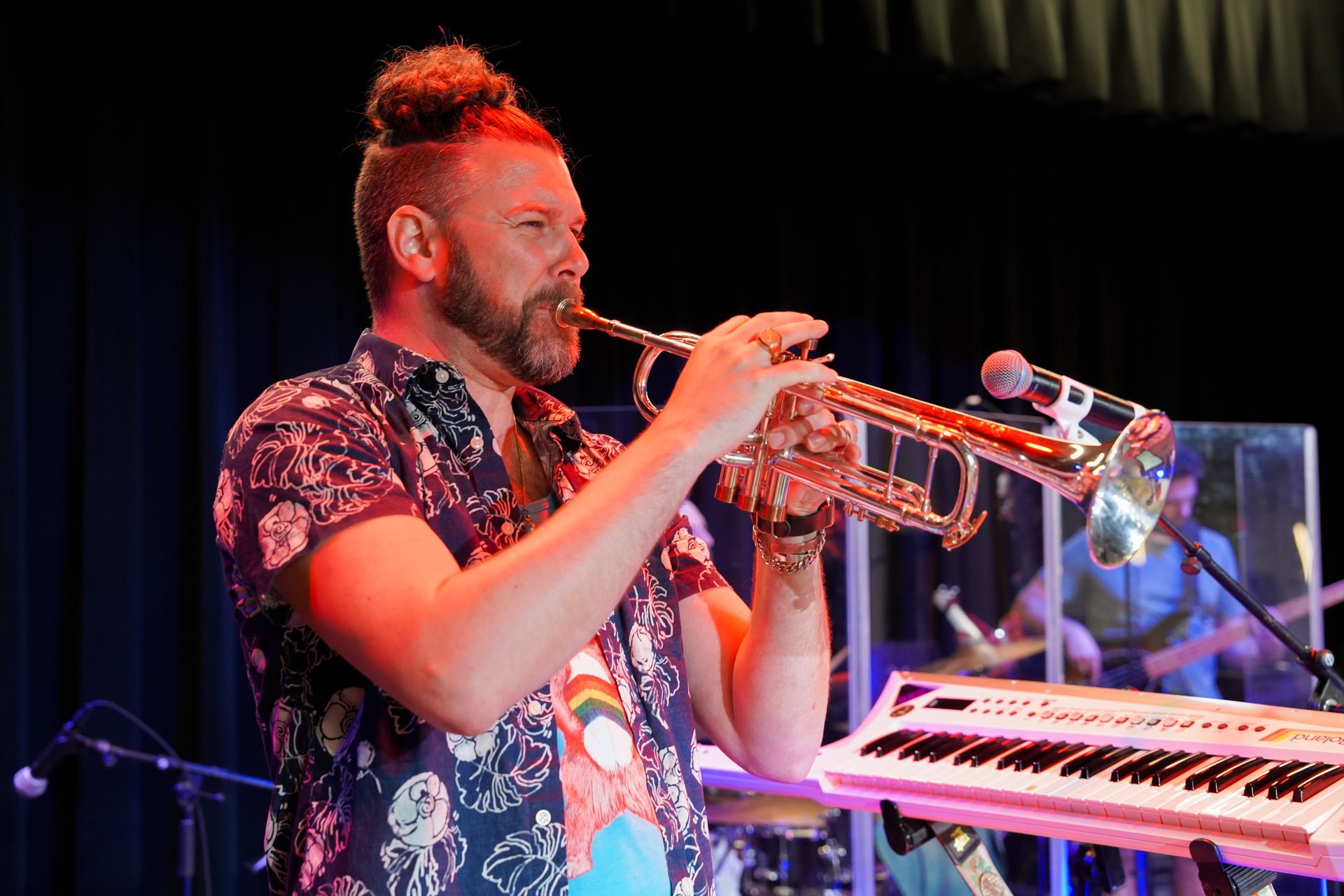 Man with topknot playing trumpet on stage, wearing a patterned shirt. Keyboard and drummer in the background.