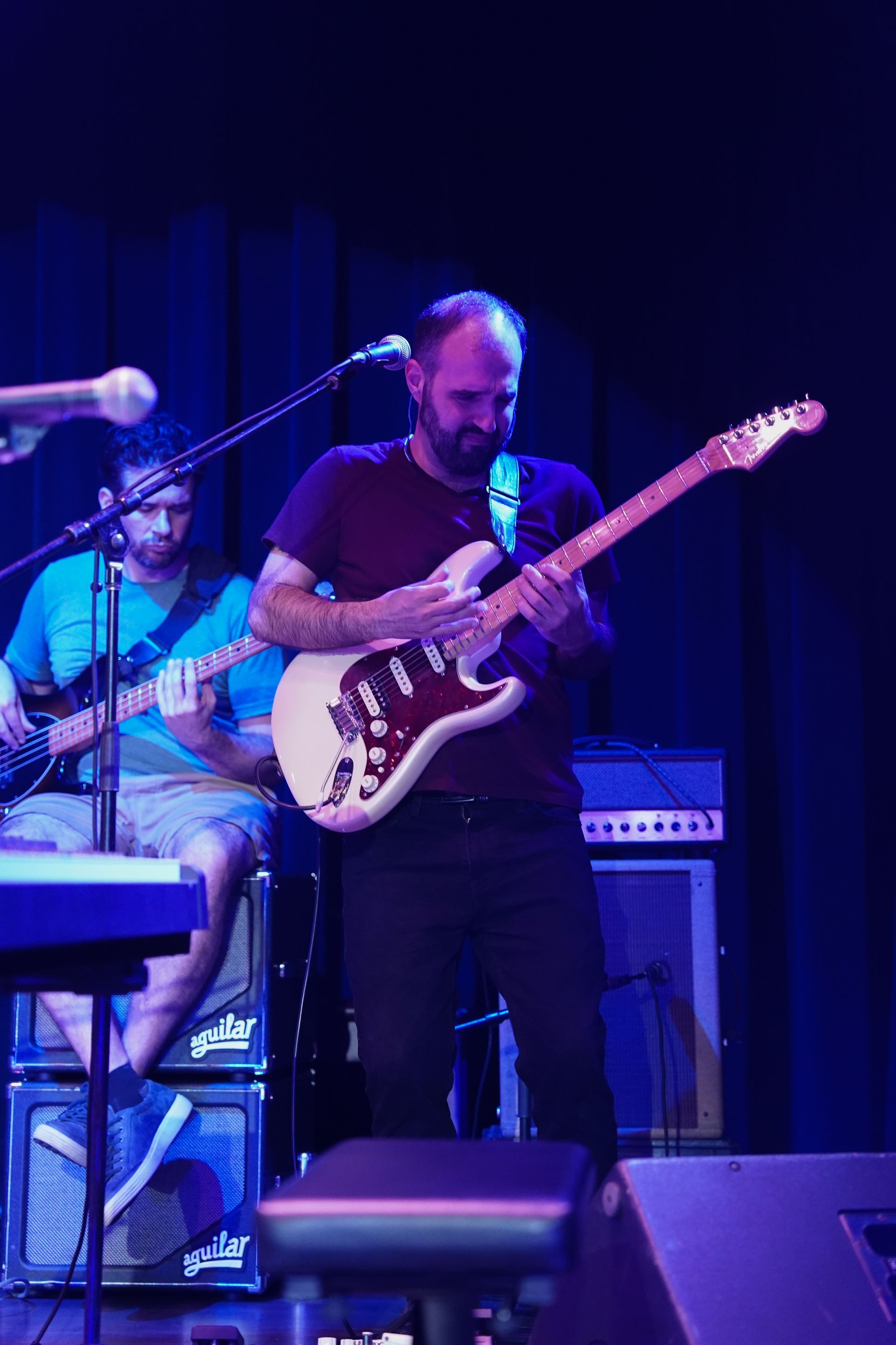 Guitarist playing on stage, wearing a dark red shirt, with another musician in the background. Blue lighting.