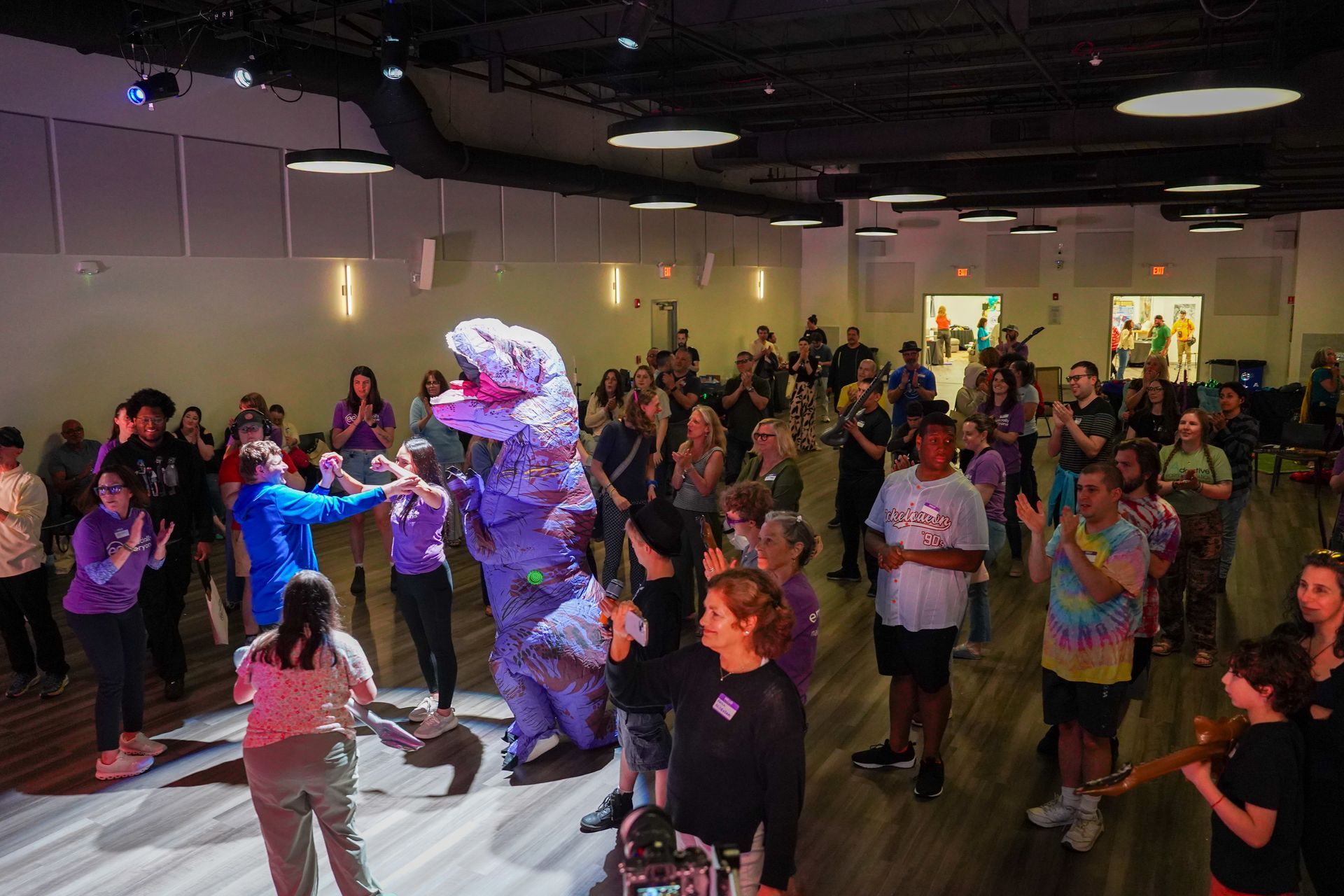 People in a large room cheer at a purple dinosaur character.