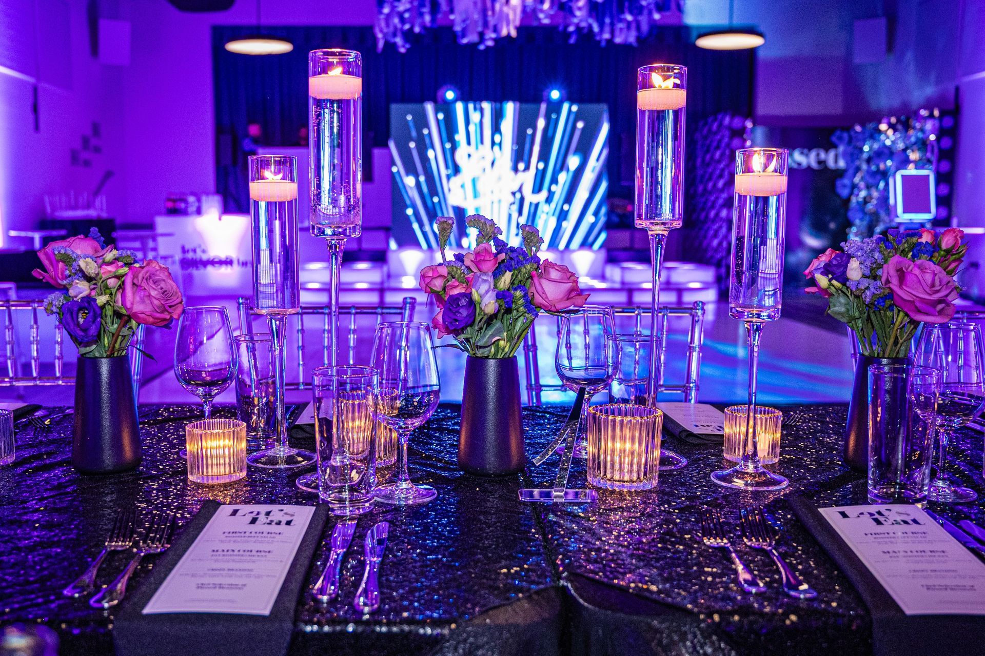 Elegant dining table with purple and gold accents, lit with candles, floral arrangements, and a screen in the background.
