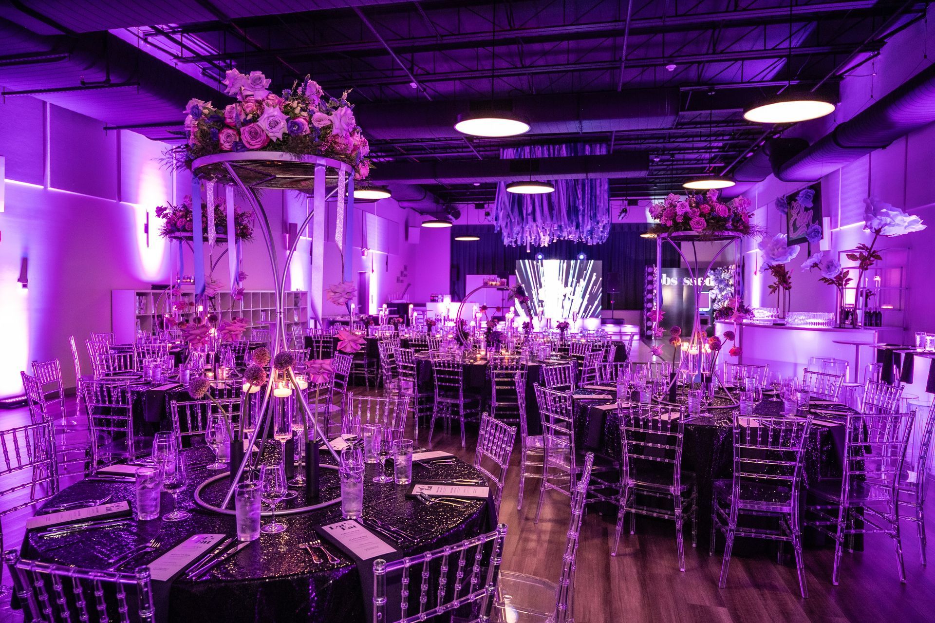 Reception hall with tables, chairs, and floral centerpieces illuminated by purple lighting.