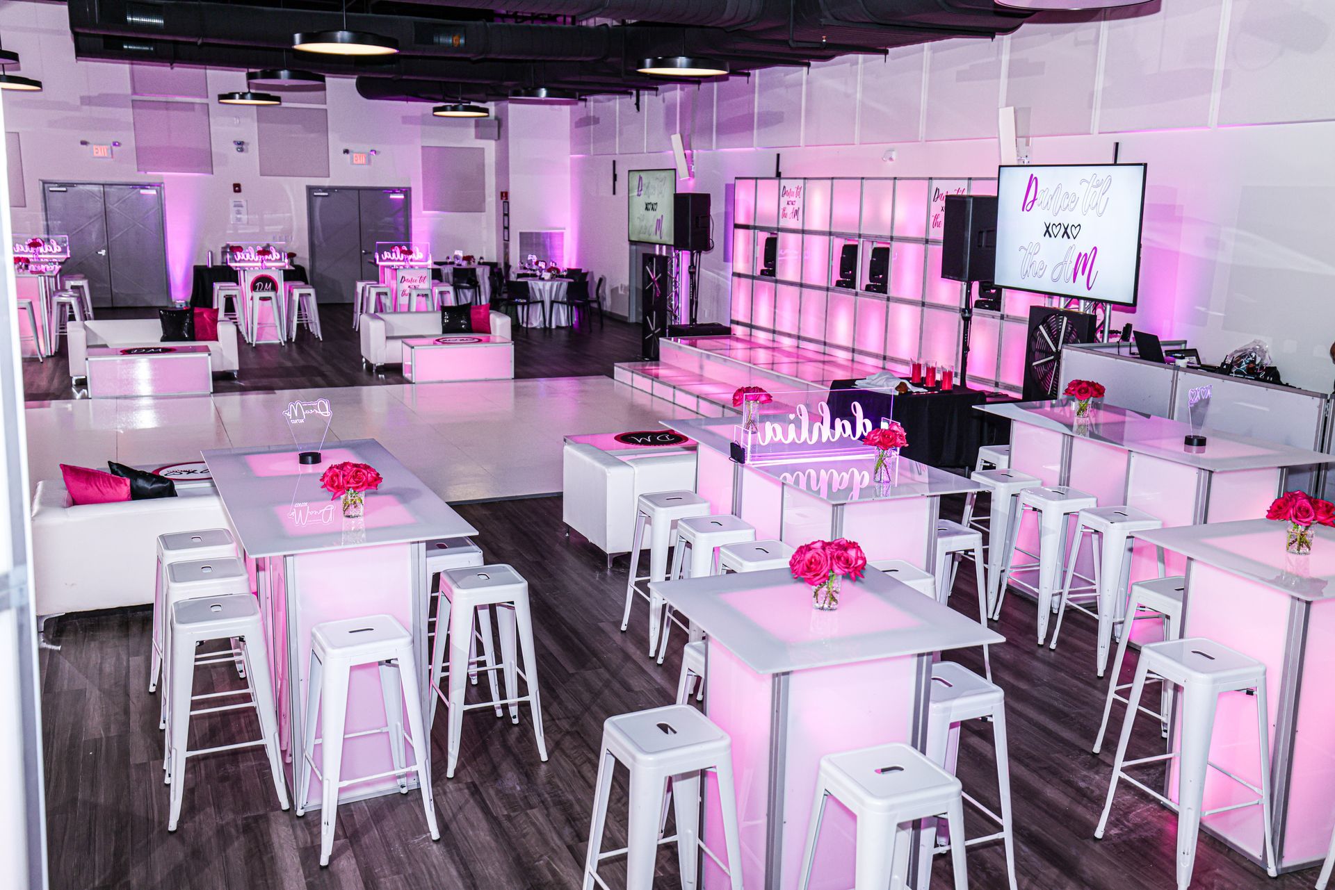 A modern event space with pink lighting, tables, white stools, and a raised stage.