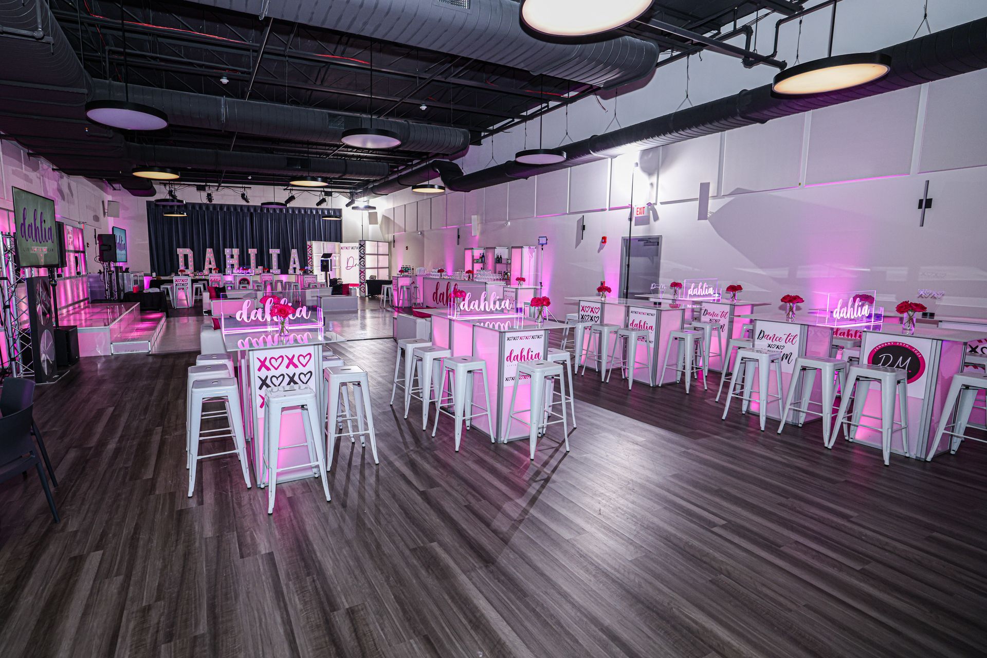 Event space with tables, chairs, and decor, illuminated by pink and white lights. Dark wood floor.