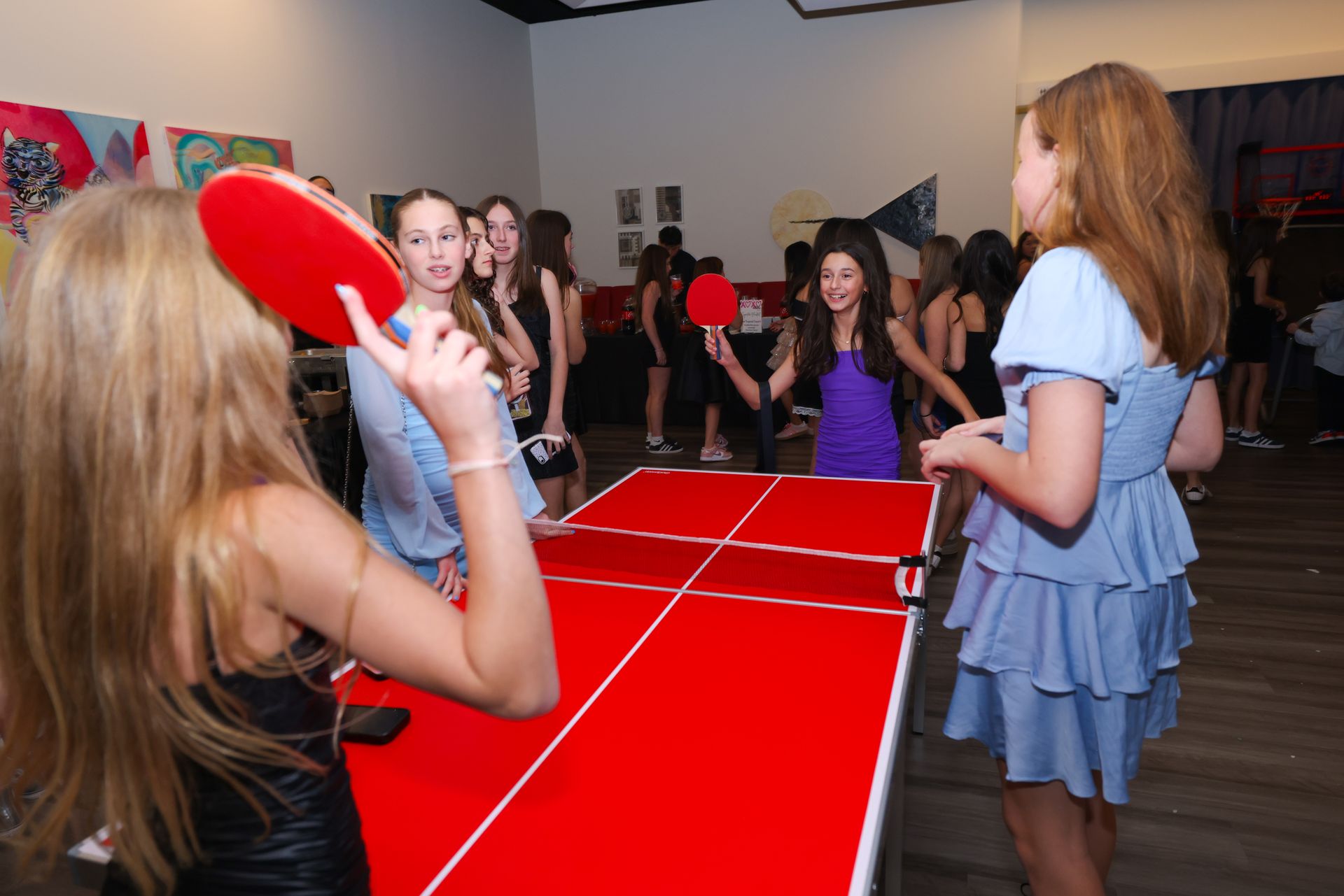 Teenagers playing ping pong at a party, red table, paddles, and onlookers.