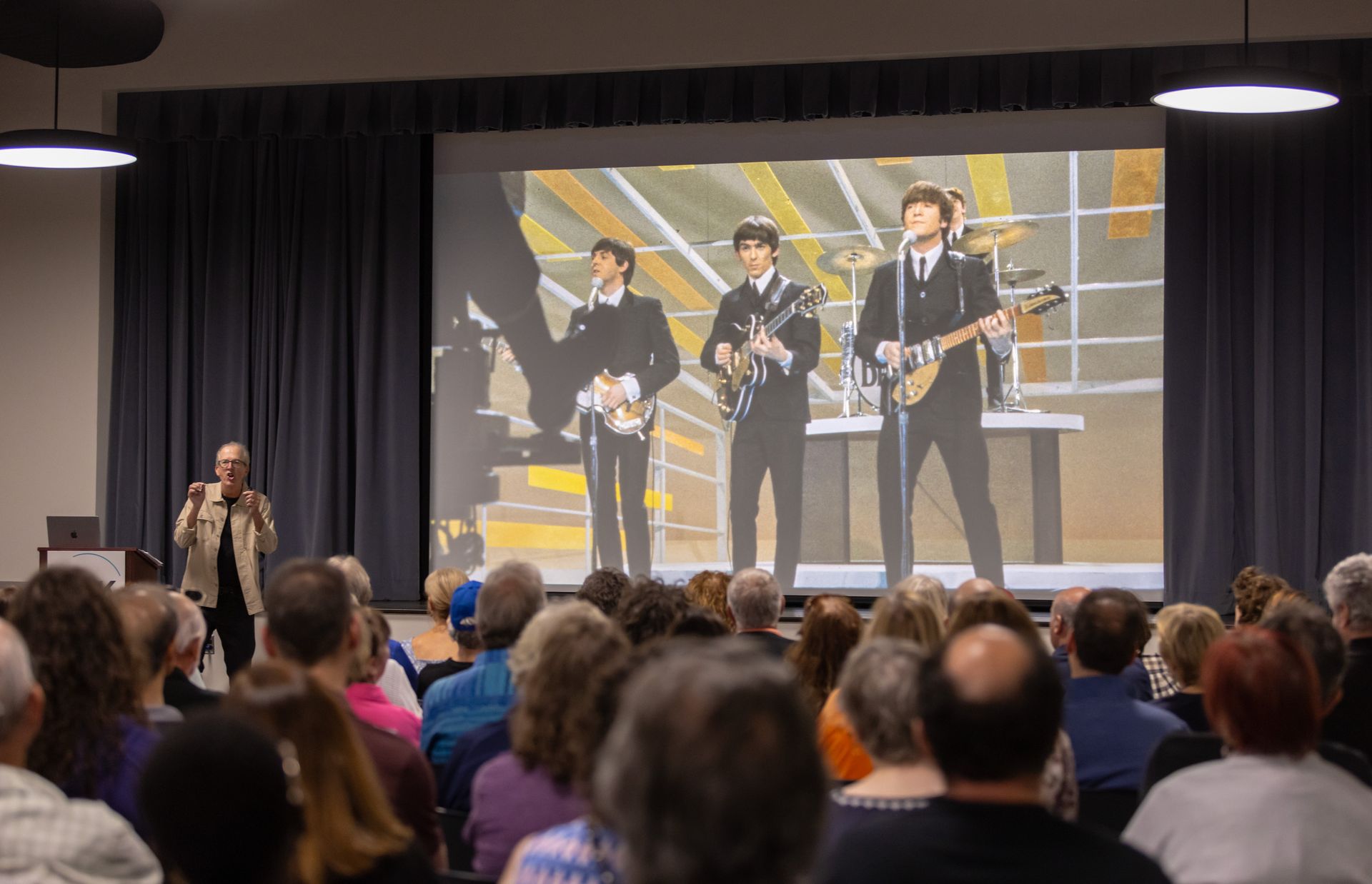 Audience watches a Beatles film projected on a screen; a speaker stands onstage.