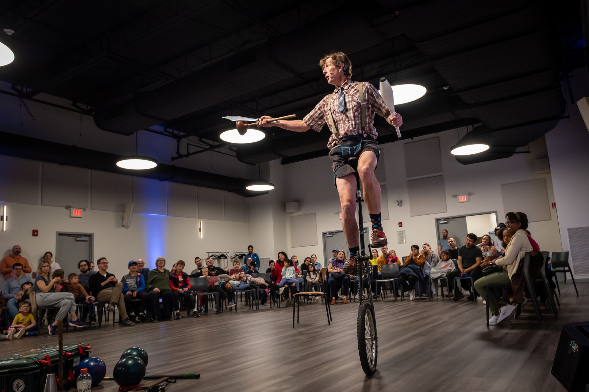 Person on unicycle juggles plates and clubs in a performance hall, crowd watches.