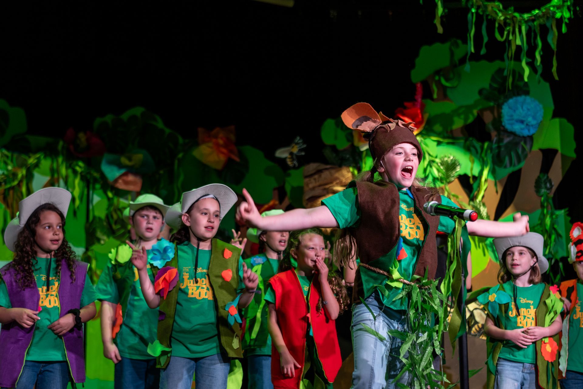 Children in green costumes performing on stage, one with a microphone, jungle backdrop.