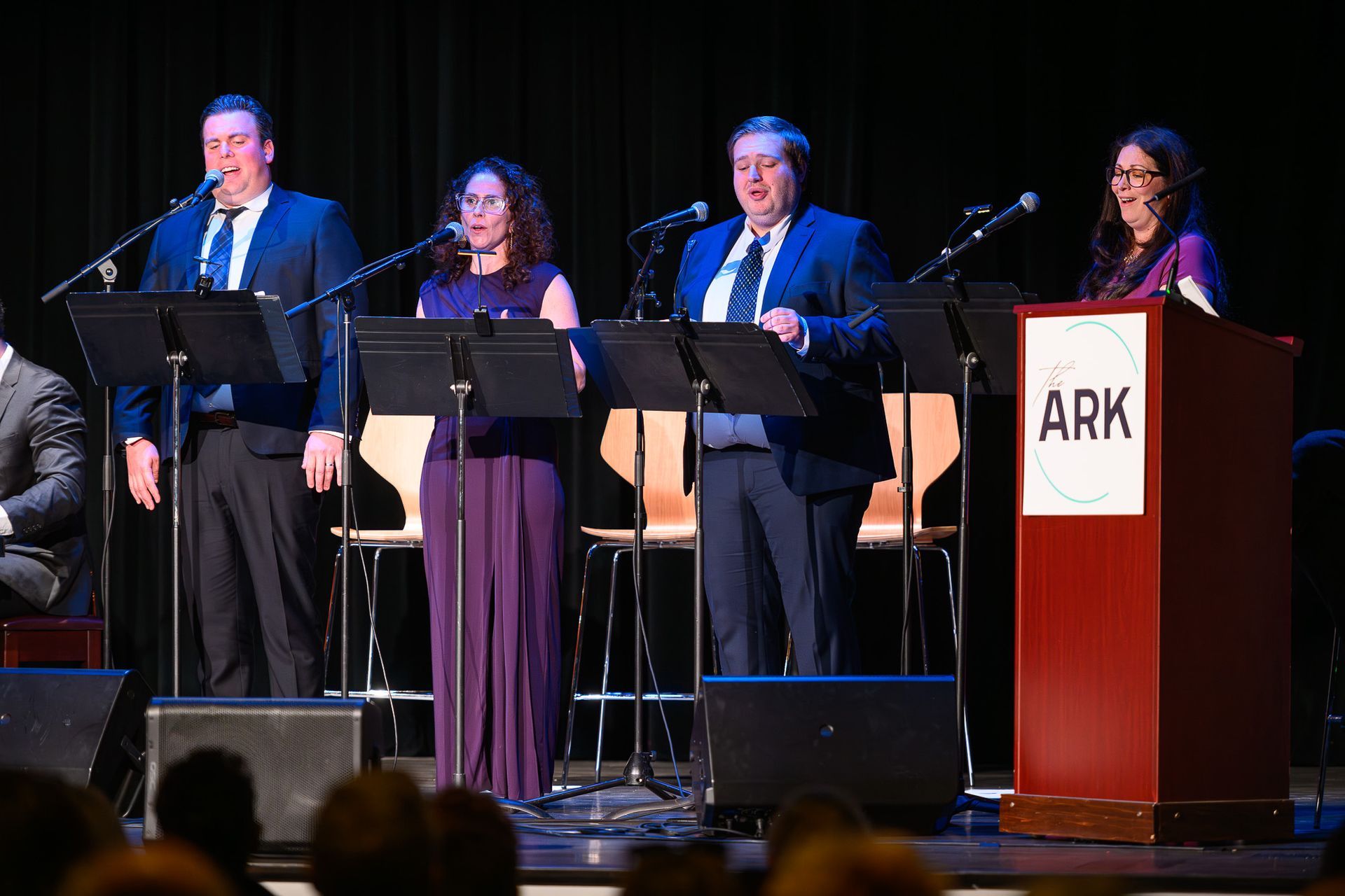 Four people singing onstage at The Ark, using music stands, wearing formal attire.