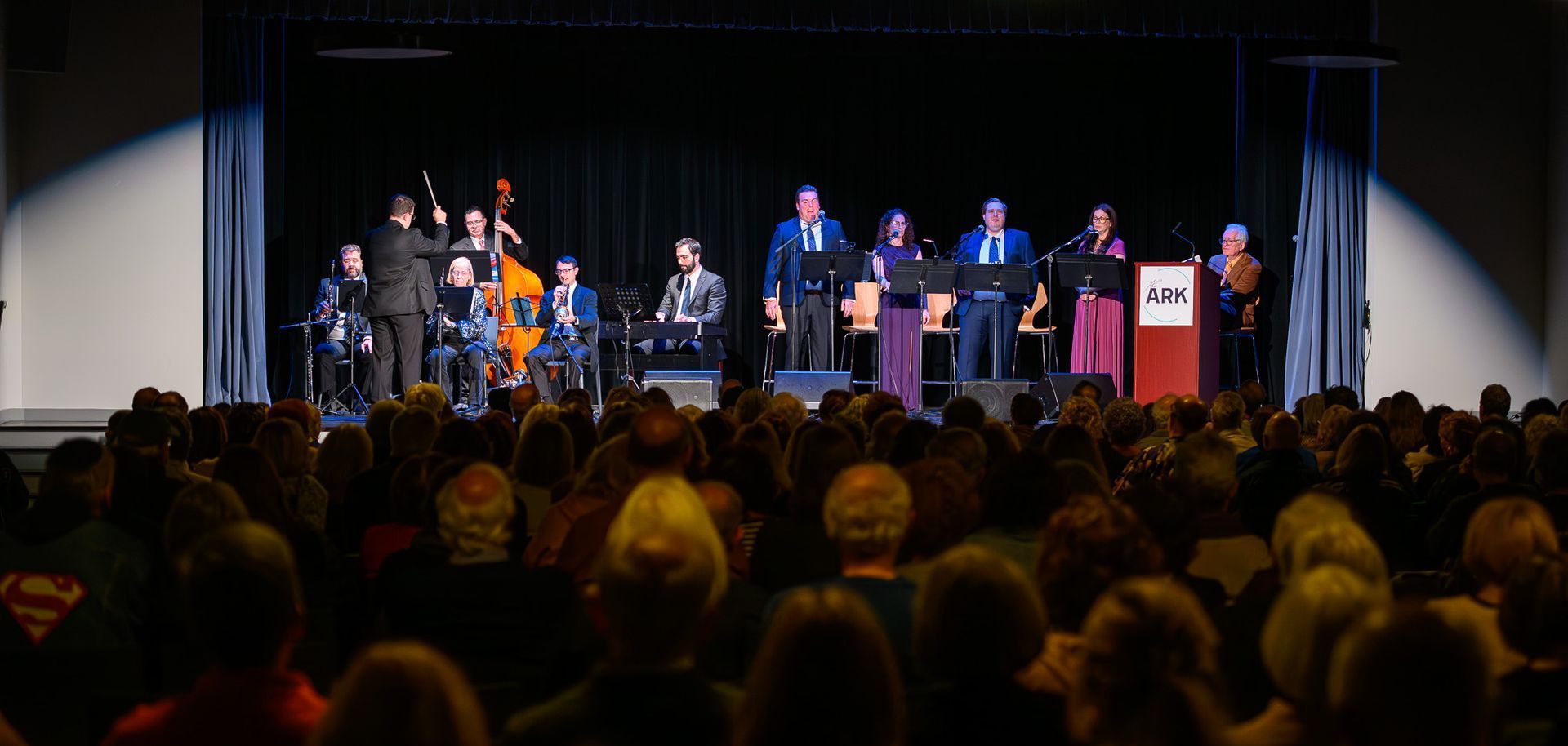 A stage performance with musicians and singers performing to a large audience seated in rows.