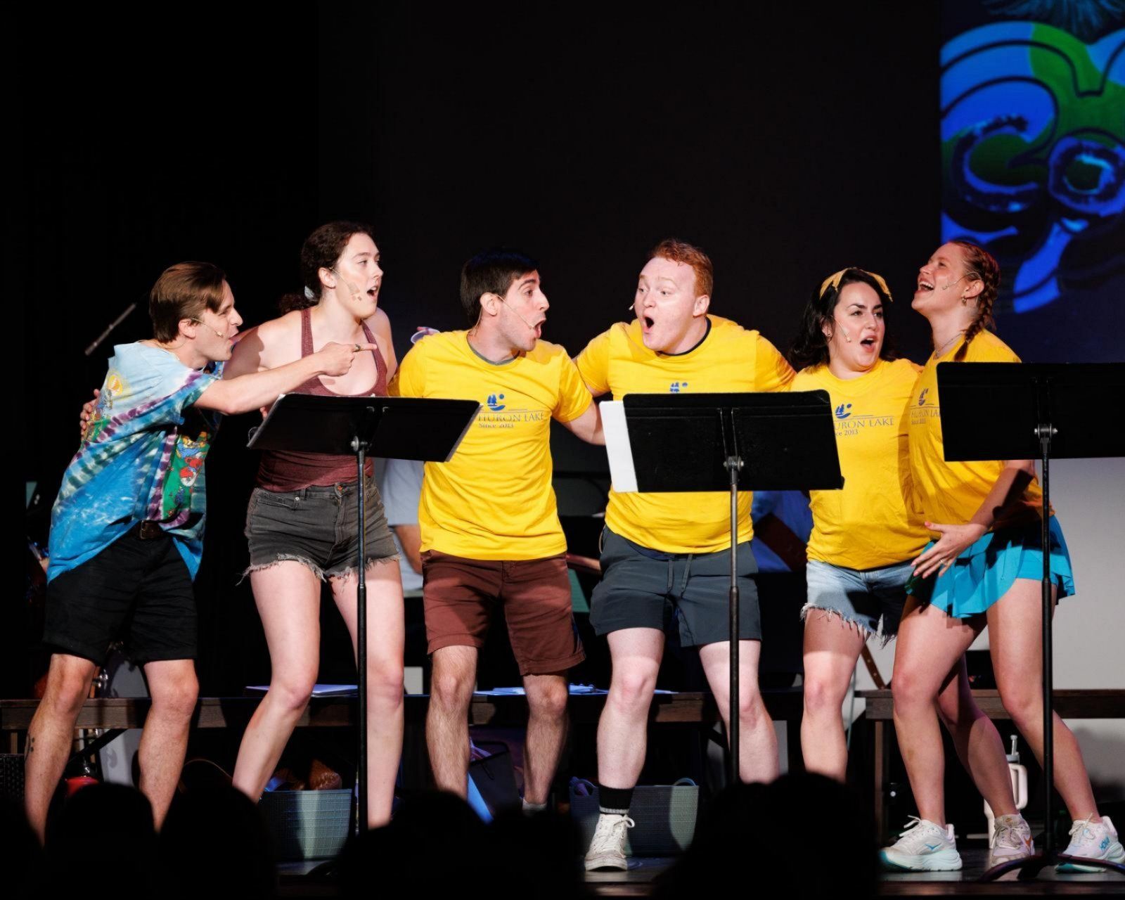 Six people on stage with surprised expressions, reading from scripts. Stage background with a blue design.