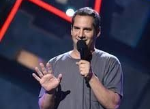 Person on stage speaking into a microphone, wearing a gray shirt and gesturing with an open hand.
