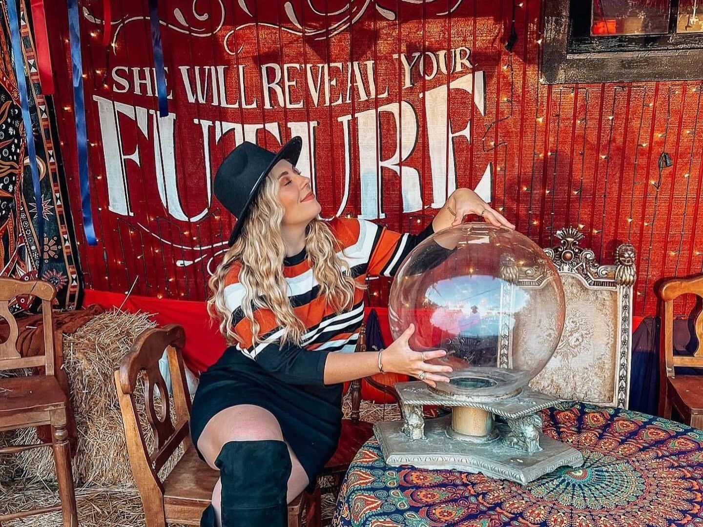 Woman in striped sweater sits beside a crystal ball in a red fortune-teller booth, with “FUTURE” sign behind.