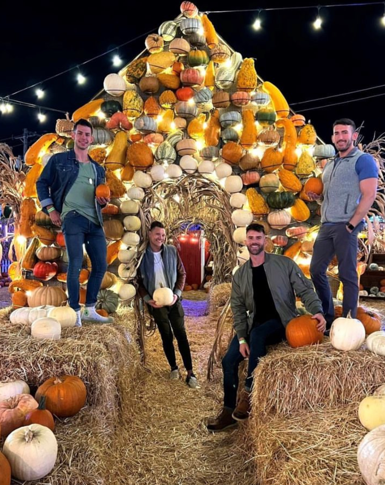 Four people posing at a pumpkin arch under string lights at a nighttime harvest display