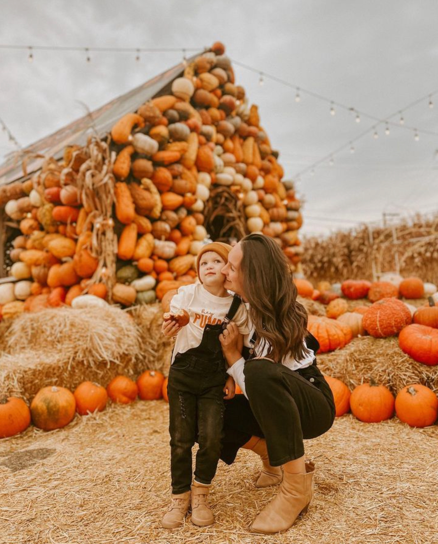 Woman and child in a pumpkin patch beside a stacked pumpkin display, with hay and string lights.