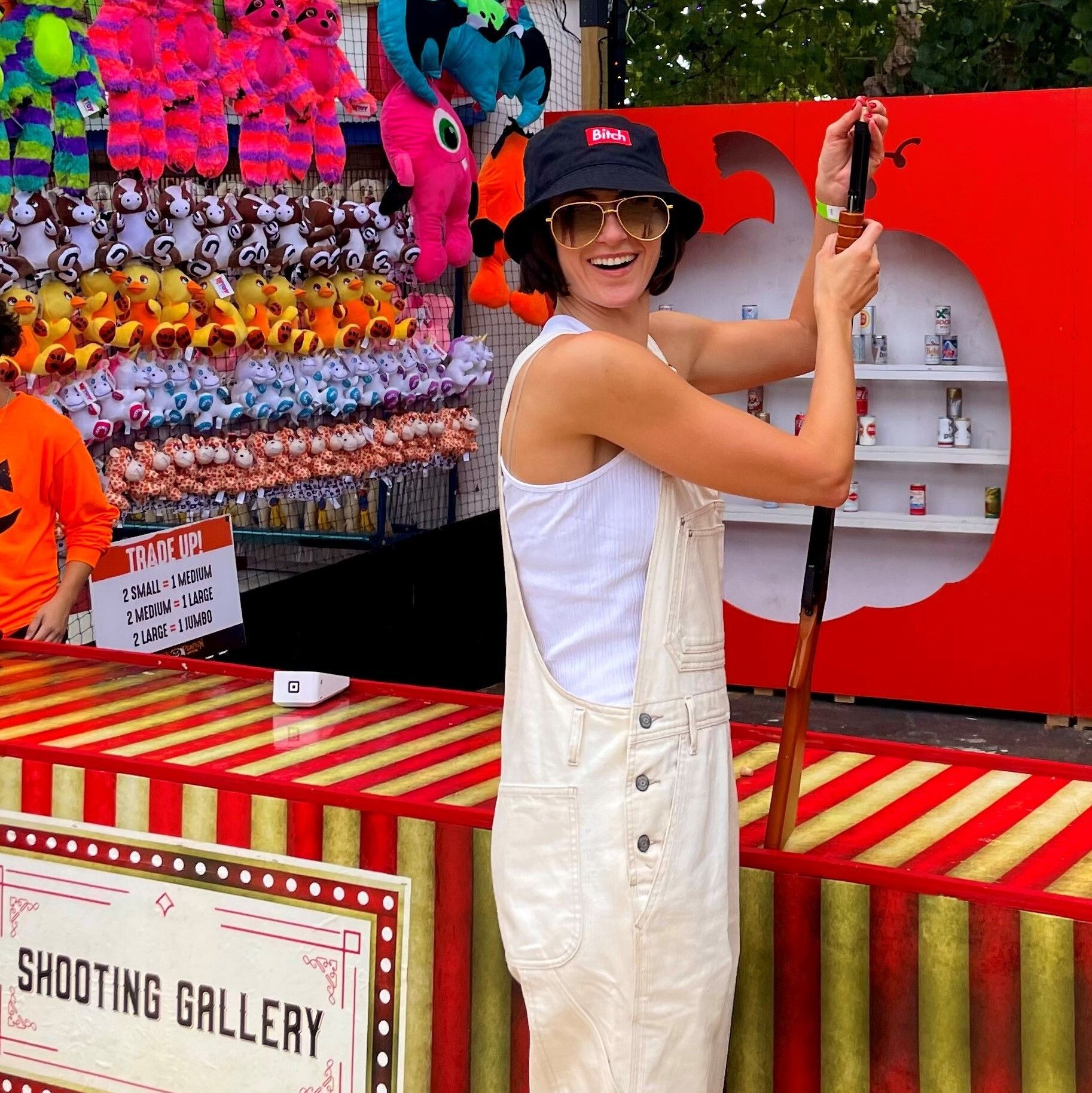Person smiling at a carnival shooting gallery, aiming a toy gun in front of colorful prizes.