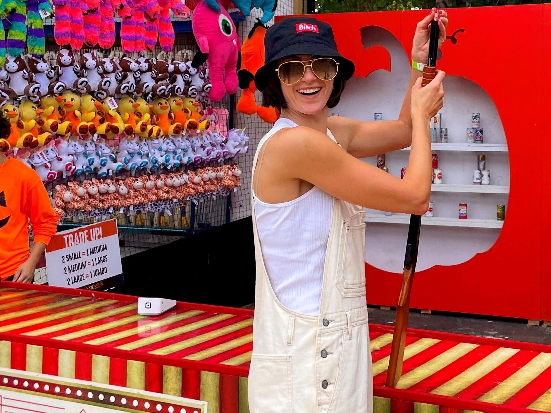 Person in white overalls and hat smiling beside a red carnival game booth with hanging prizes