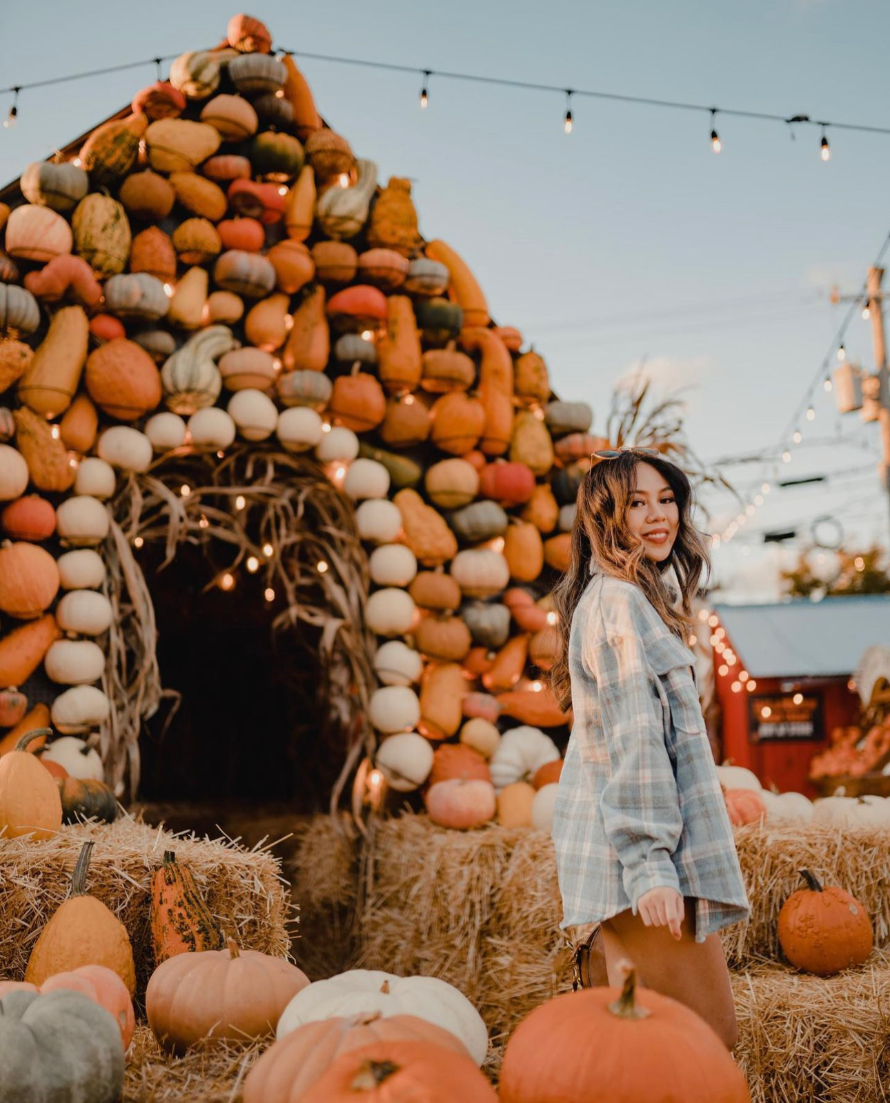 Woman standing beside a pumpkin-covered hay bale arch at an autumn festival at dusk