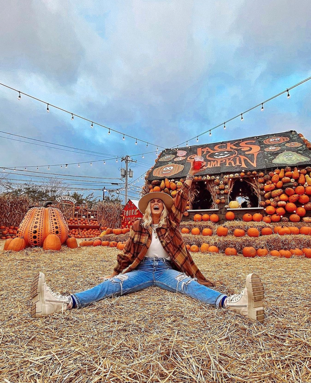 Person sitting in a hay-covered pumpkin patch, flanked by giant pumpkins and a decorated pumpkin stand.