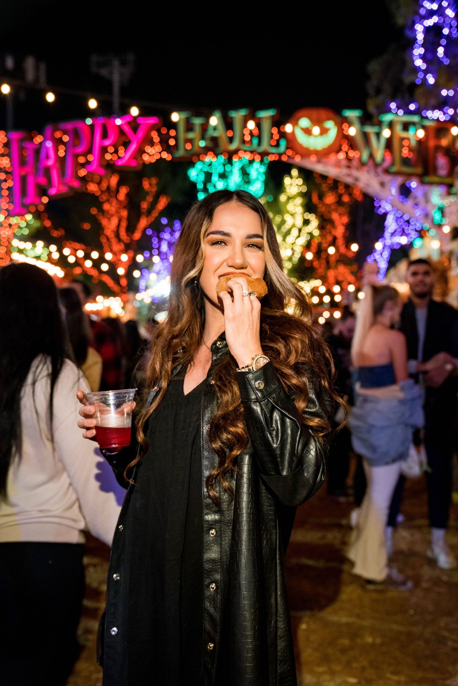 Woman in black leather jacket smiling at a Halloween party under colorful lights and a “Happy Halloween” arch