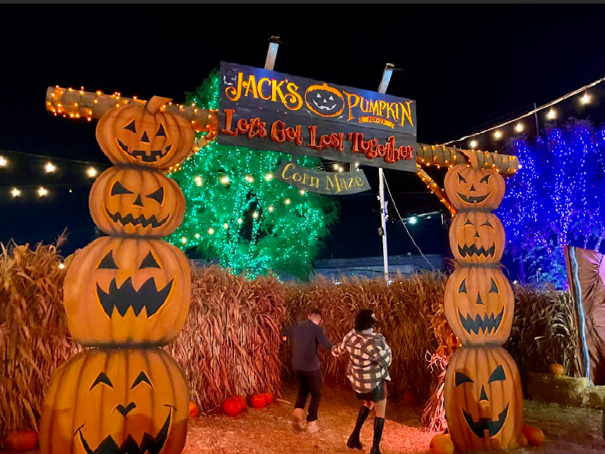 Halloween entrance with stacked pumpkin totems, lit sign, hay bales, and a child walking under colorful lights